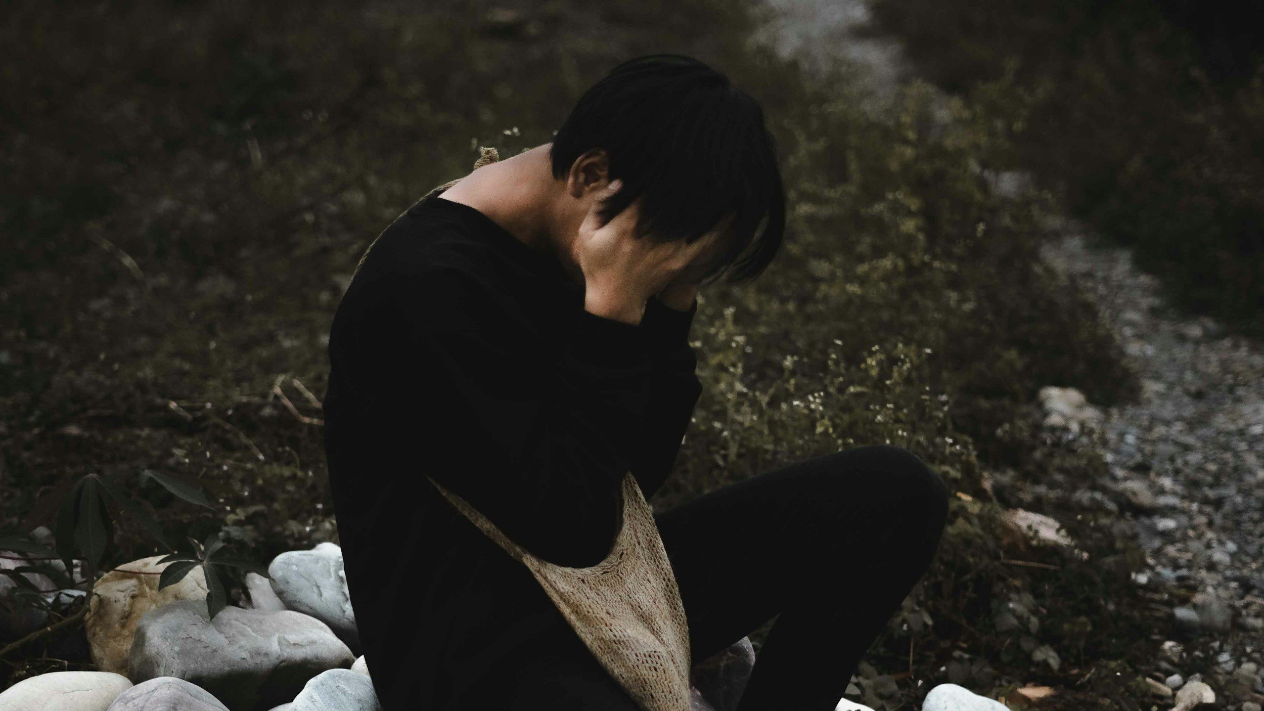Person sitting on rocks on the ground, covering their face with their hands, in a dark outdoor setting.