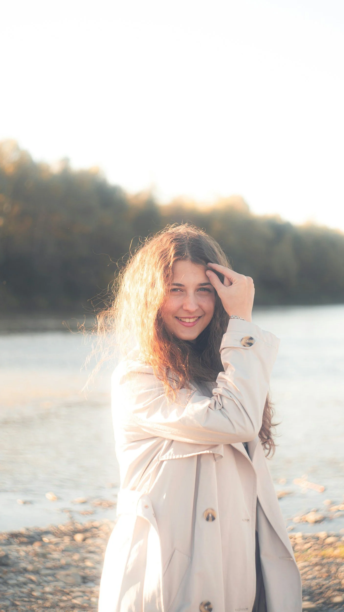 A woman with curly hair smiling on a beach during sunset, wearing a beige trench coat.