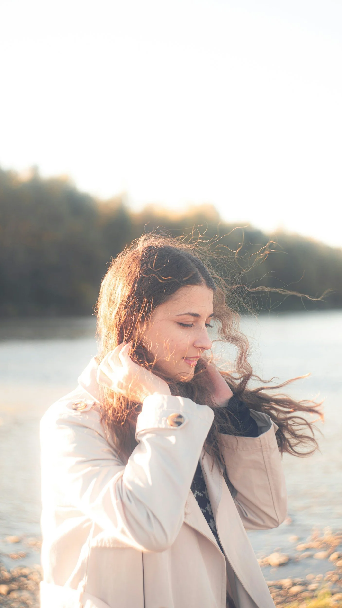 A woman with long curly hair, wearing a beige coat, is outdoors near a body of water during sunset, with trees in the background, her eyes closed and a peaceful expression.