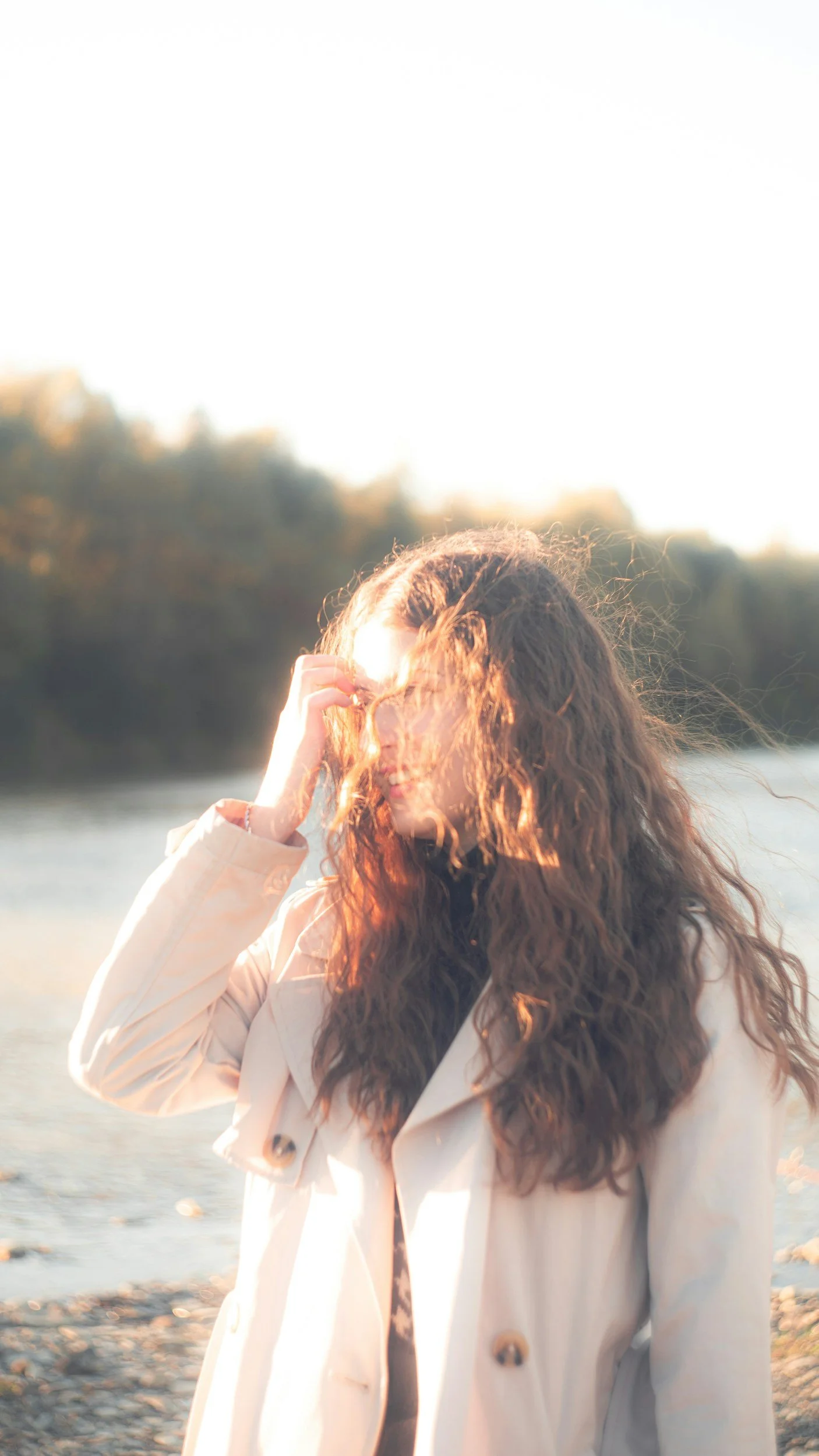 A woman with curly hair wearing a beige trench coat standing outdoors near water, with trees in the background, illuminated by bright sunlight.