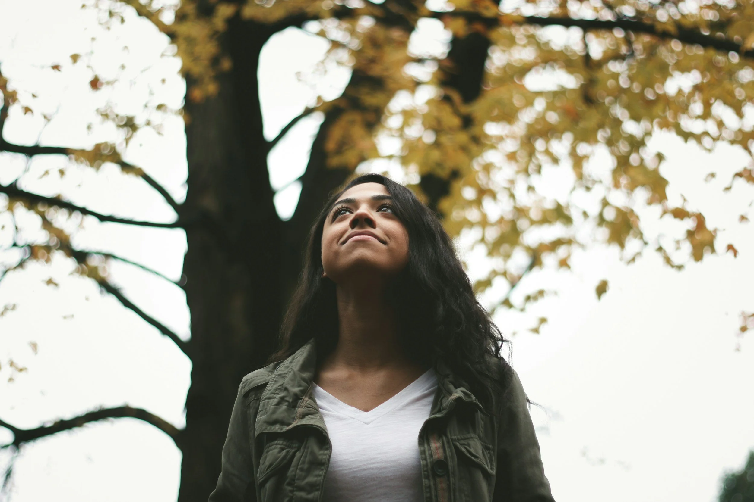 A woman with long dark hair standing outdoors under a tree with yellowing leaves, looking up with a slight smile, wearing a white shirt and a green jacket.