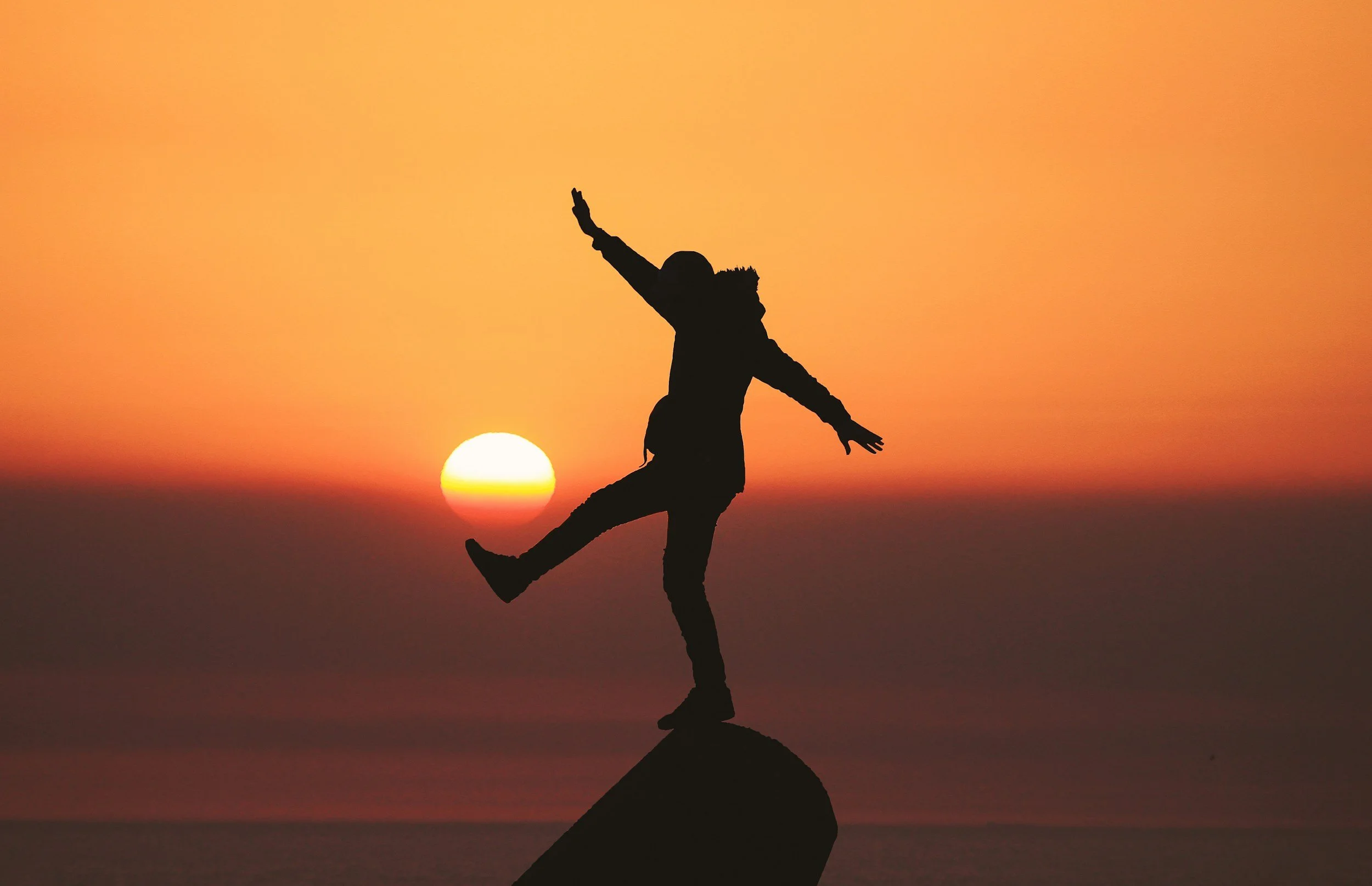 Person standing on a rock at sunset, with arms raised, silhouetted against an orange sky and the setting sun.