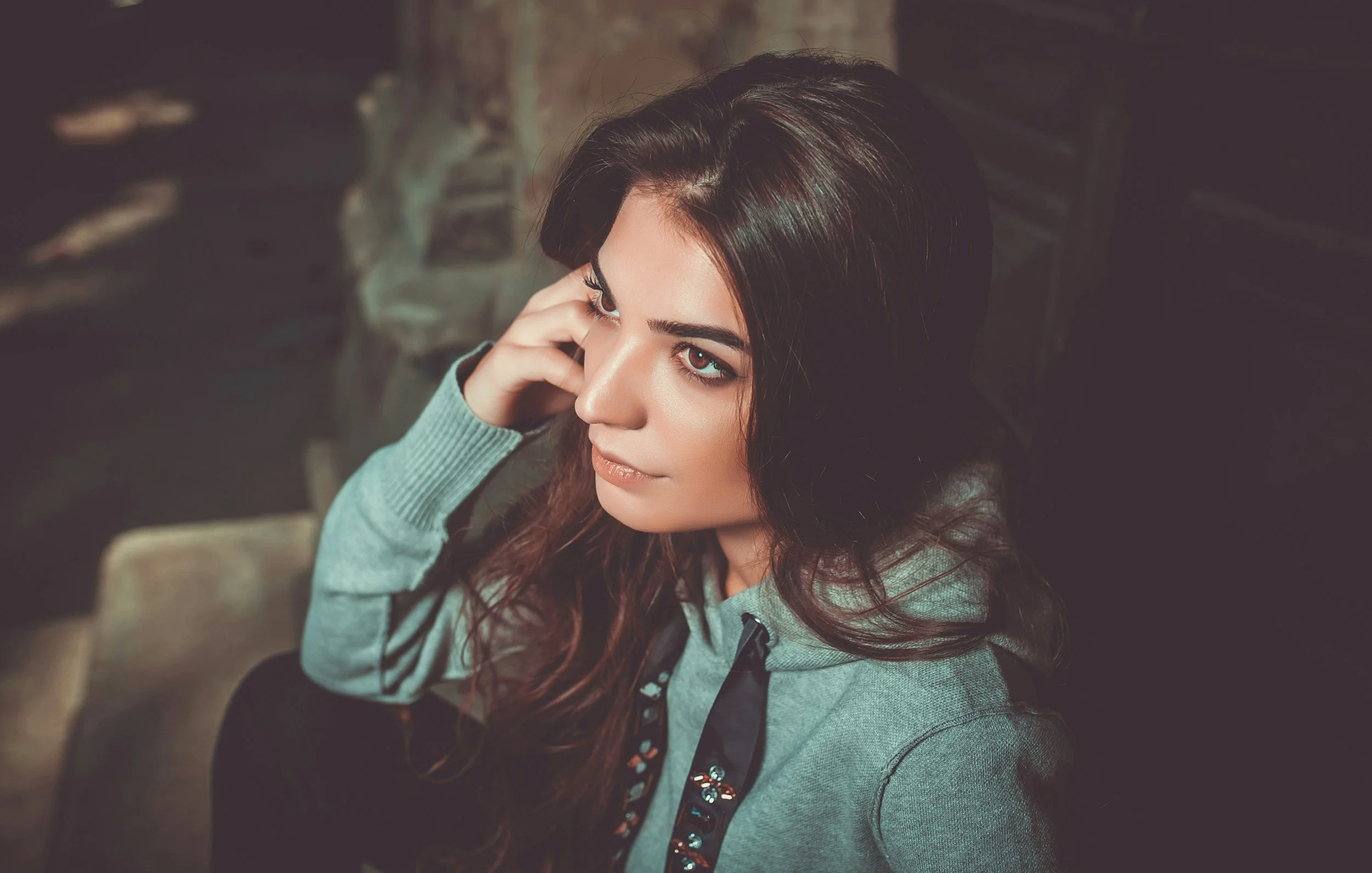 A young woman with long dark hair and bright eyes looks up thoughtfully, resting her hand on her face, wearing a gray hoodie with black accents.