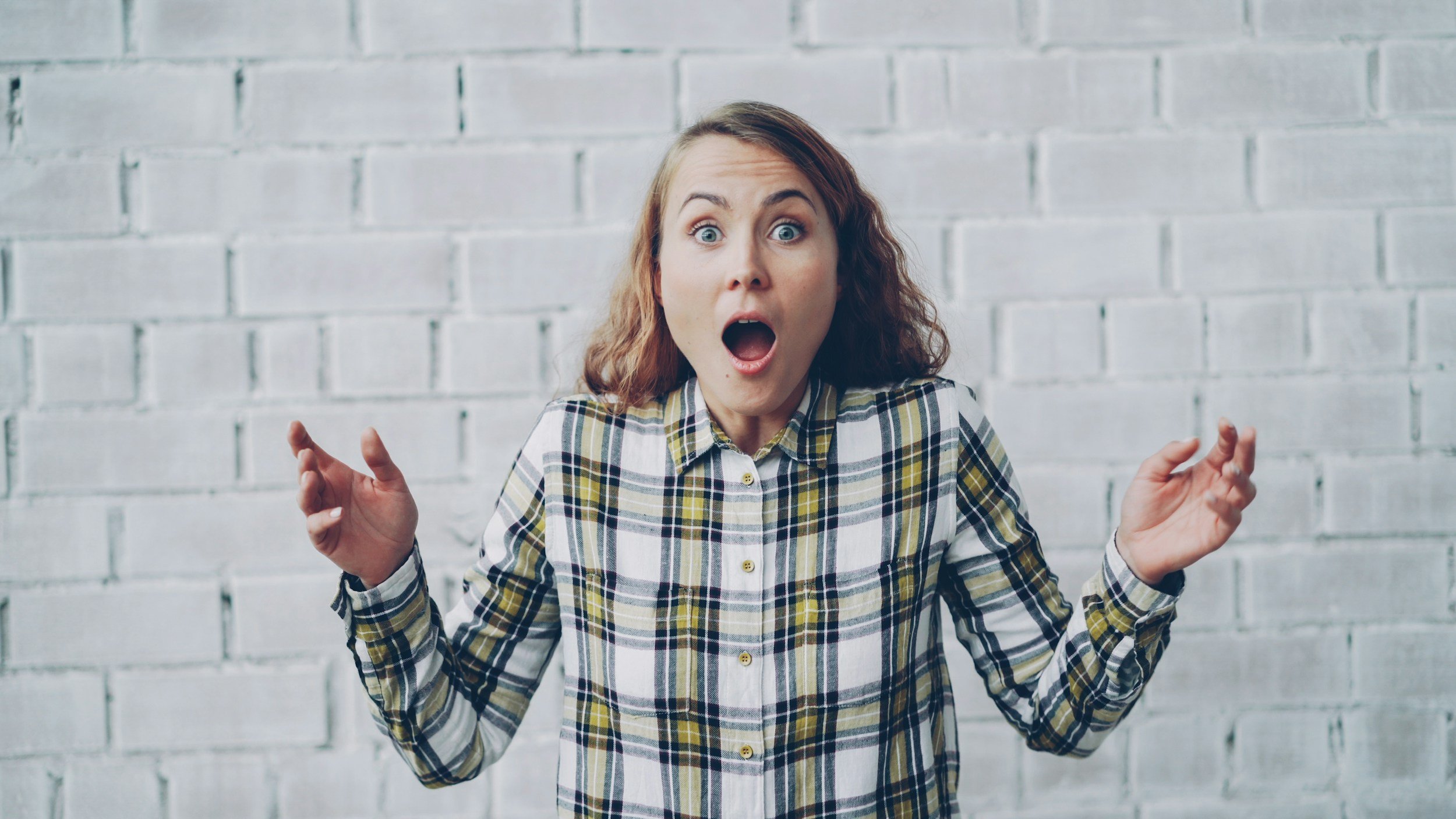 A women with her mother ajar and her hands up signaling overwhelm. She is wearing a plaid white, black and yellow flannel shirt with long sleeves.