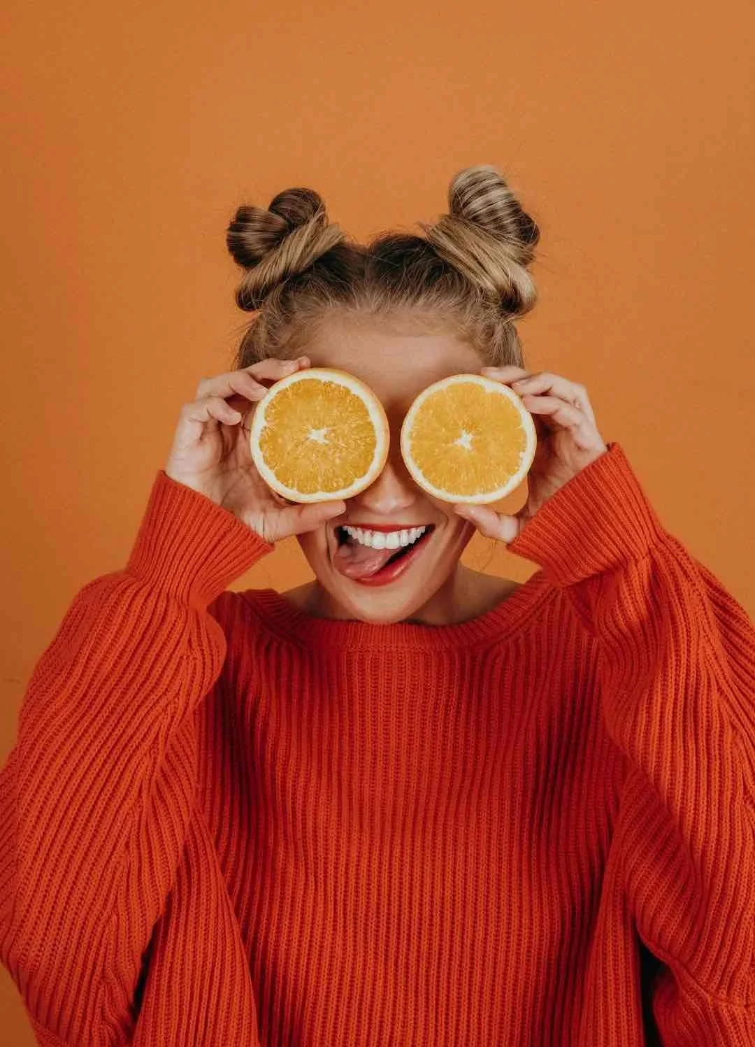 A woman with light brown hair styled in two buns, wearing an orange sweater, holding sliced oranges over her eyes, sticking her tongue out playfully against an orange background.