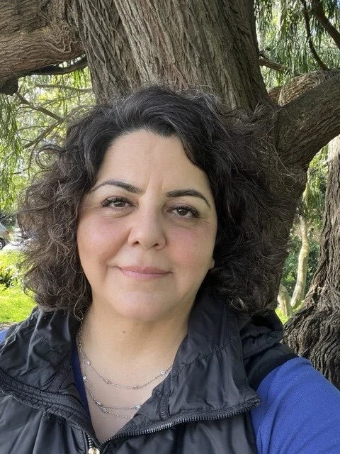 A woman with curly dark hair and fair skin taking a selfie outdoors in front of a large tree with green leaves.