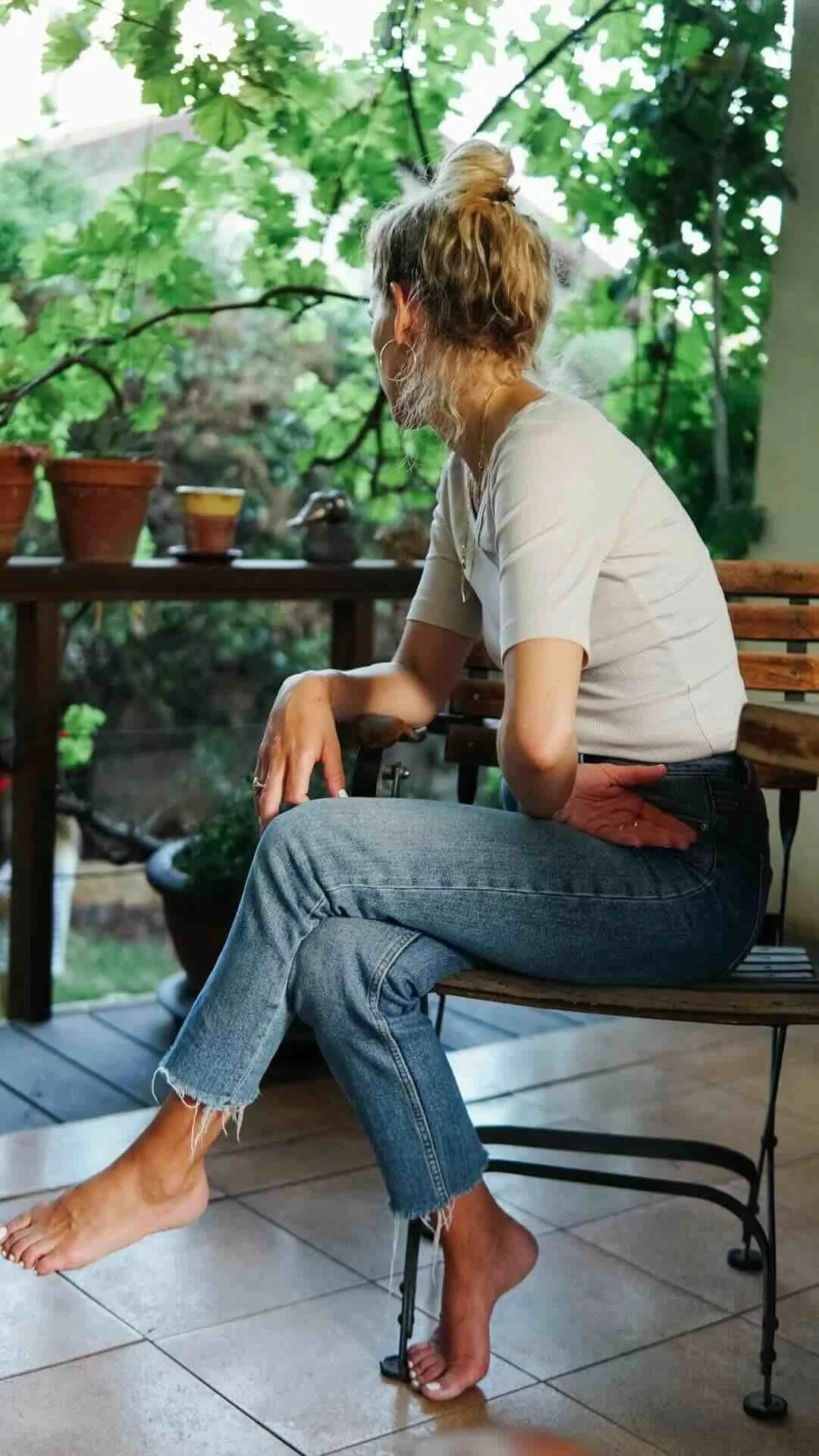 A woman sitting on a wooden bench on a porch, with one foot resting on a stiletto heel and the other foot on the floor. She is looking out at a leafy green backyard, holding a cigarette in her right hand. She has curly hair tied in a bun, is wearing a beige top and ripped blue jeans.