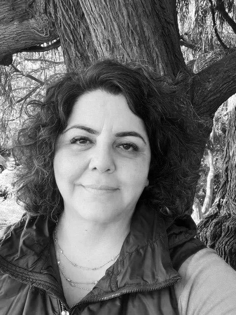 Black and white photo of a woman with curly hair smiling in front of a large tree.