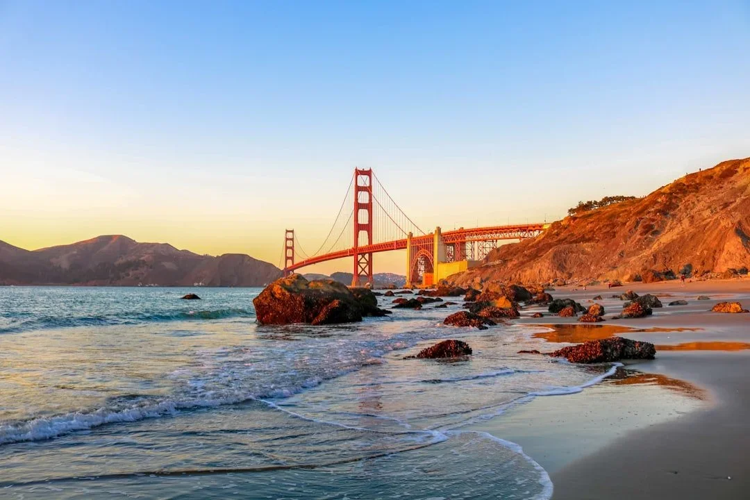 Golden Gate Bridge over the ocean at sunrise, with rocks and sandy beach in foreground and hills in background.