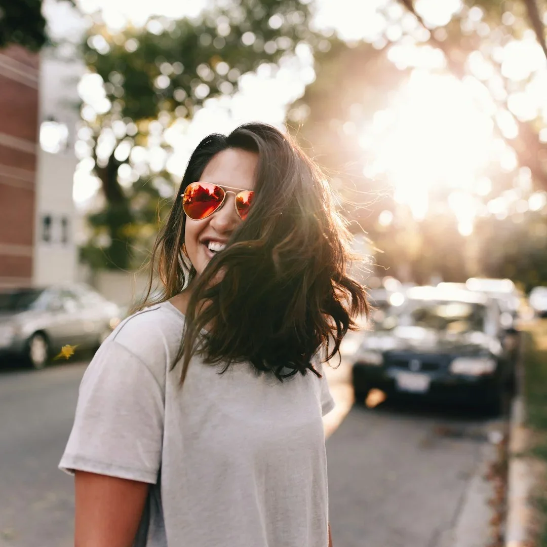 A smiling woman with wavy hair wearing sunglasses walking outdoors during sunset, with cars and trees in the background.