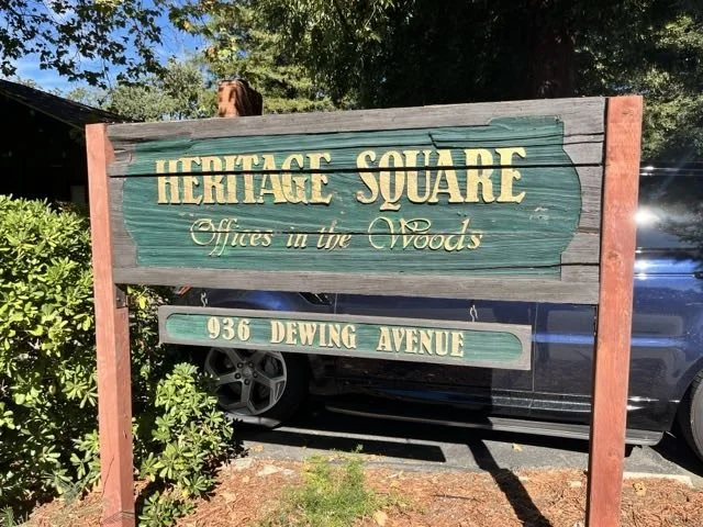 A wooden signboard with green paint and cream-colored text, reading 'Heritage Square Offices in the Woods' and the address '036 Dewing Avenue,' placed outdoors next to bushes and a parking area with cars.