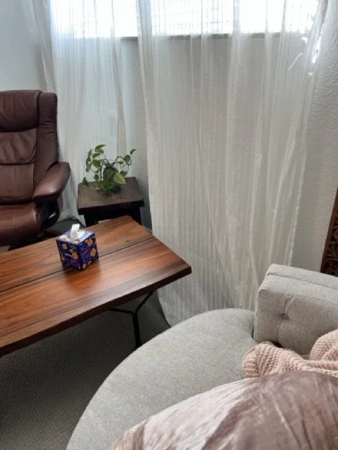 Therapy room with a wooden table, a tissue box, a beige armchair with a pink knitted blanket, a brown leather armchair, a small side table with a plant, sheer white curtains, a window with horizontal blinds, a wall clock, and textured beige walls.