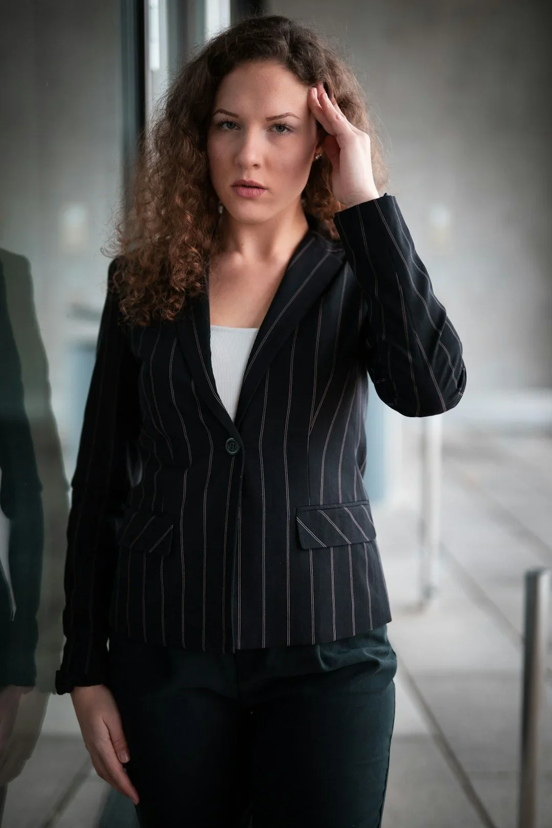 A woman with curly brown hair and a serious expression, wearing a black pinstripe blazer over a white top, standing indoors near a glass wall.