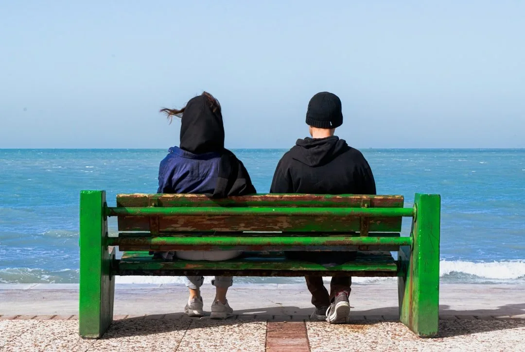Two people sitting on a green bench facing the ocean, with a clear blue sky