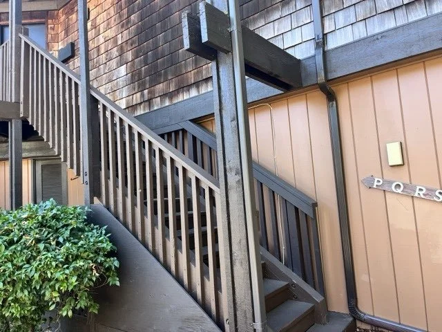 Outdoor wooden staircase with handrails leading to an upper level, adjacent to a brown wooden building, with a bush in the foreground and a wall-mounted directional sign to the right.