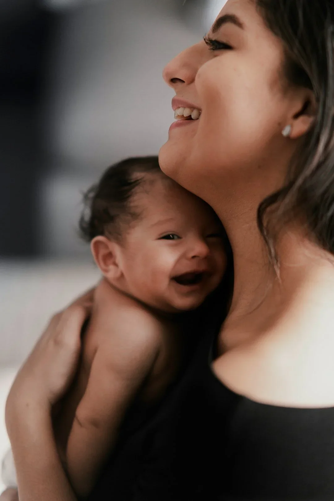 A woman with dark hair smiling and holding a smiling baby close to her chest.