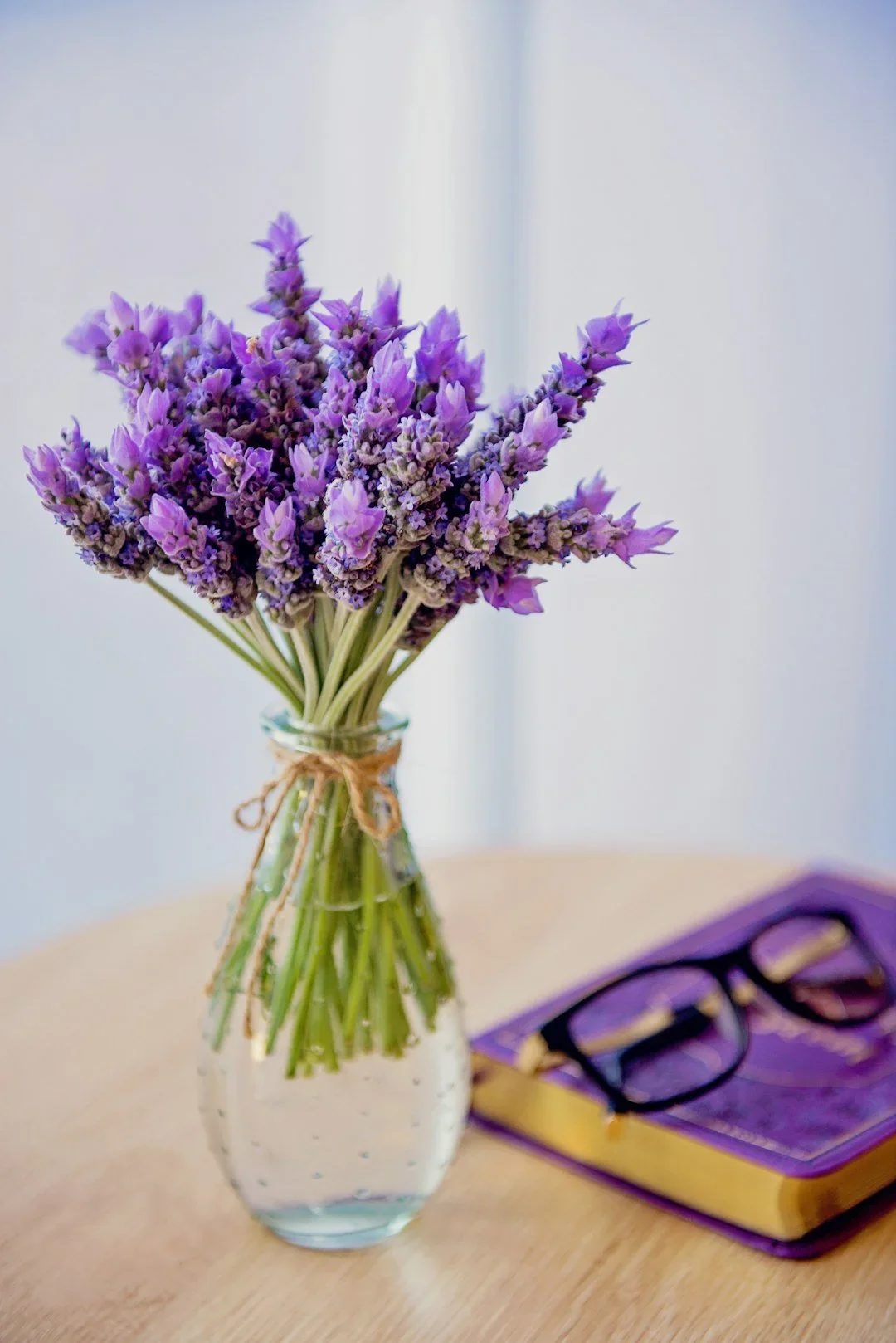 A lavendar bouquet in a vase. A lavendar colored book beside it on the table with black reading glasses ontop.