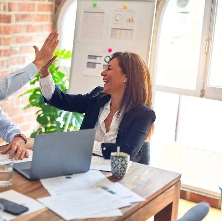 Two colleagues high-fiving in a bright office, with a laptop and papers on the desk, and a whiteboard with charts in the background.