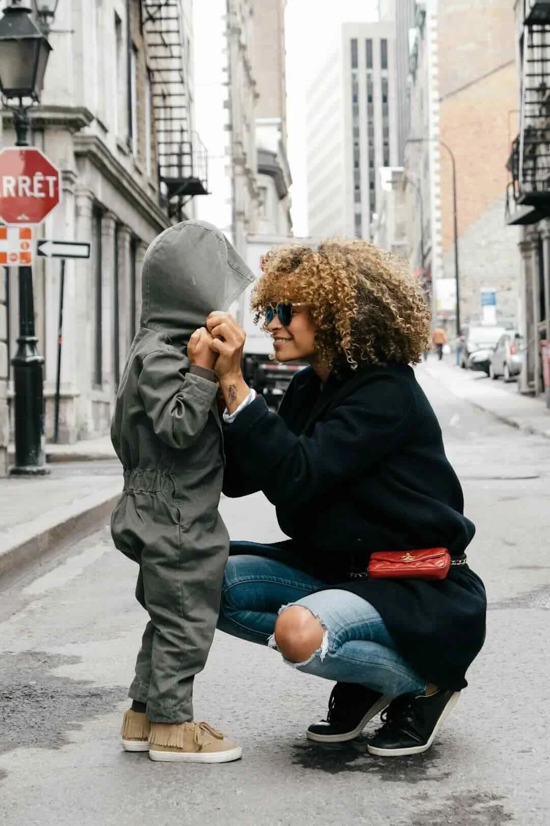 A woman with curly hair wearing sunglasses, a black coat, and ripped jeans squats on the street while holding hands with a young child dressed in a gray hooded jumpsuit. The woman is smiling and engaging with the child in an urban setting with buildings, parked cars, and street signs visible in the background.