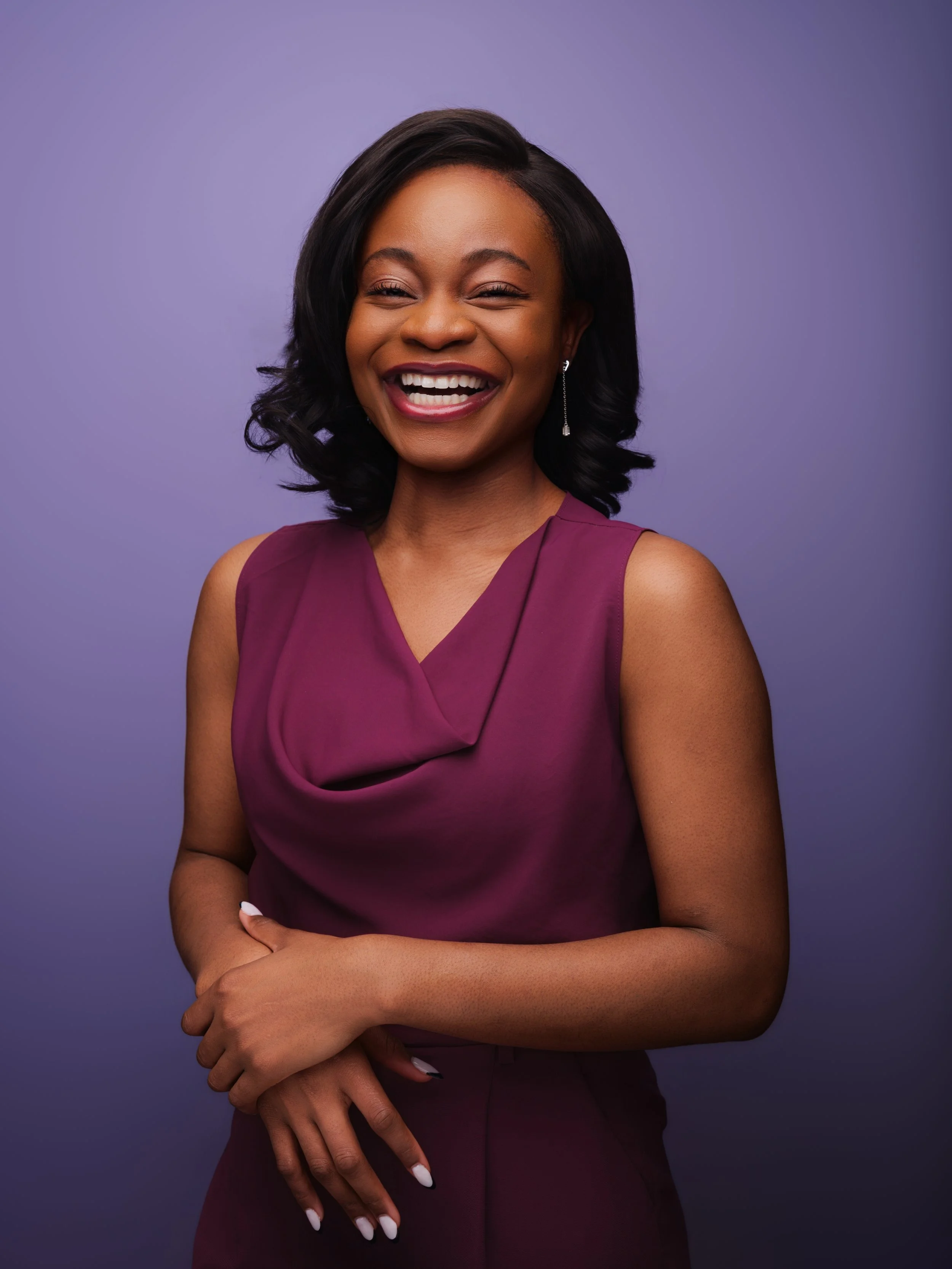 A woman with dark hair styled in loose waves, wearing a sleeveless burgundy dress, standing against a purple background, smiling broadly with arms crossed.