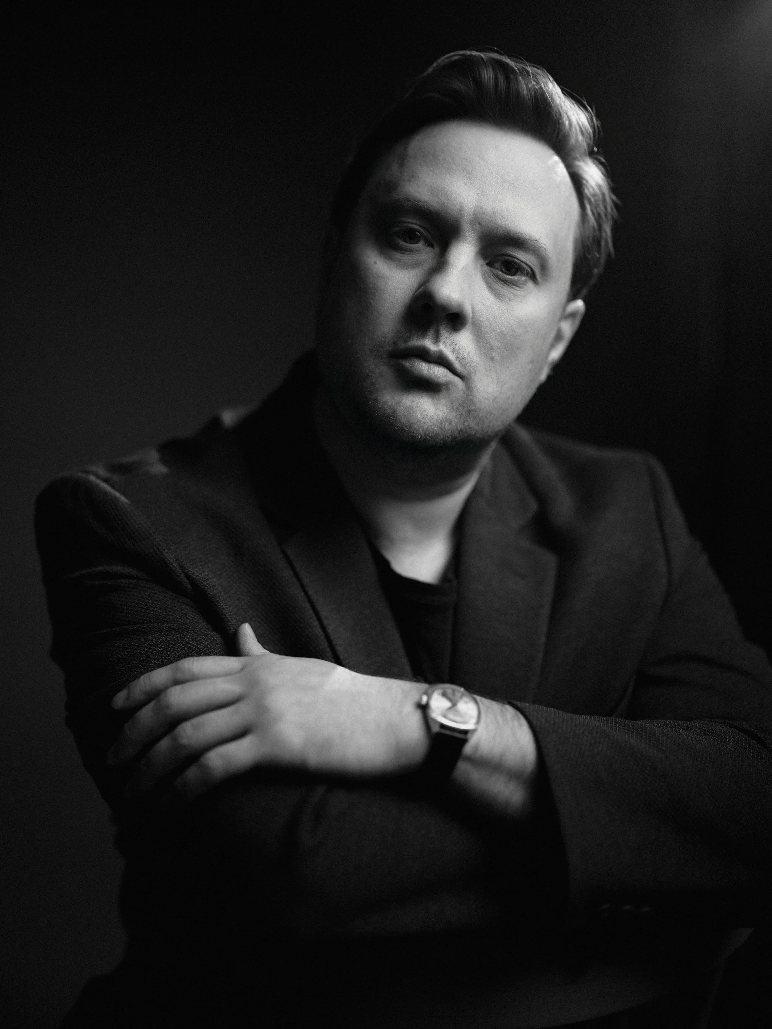 Black and white portrait of a man with short dark hair, wearing a dark blazer and a watch, looking at the camera with a serious expression, against a dark background.