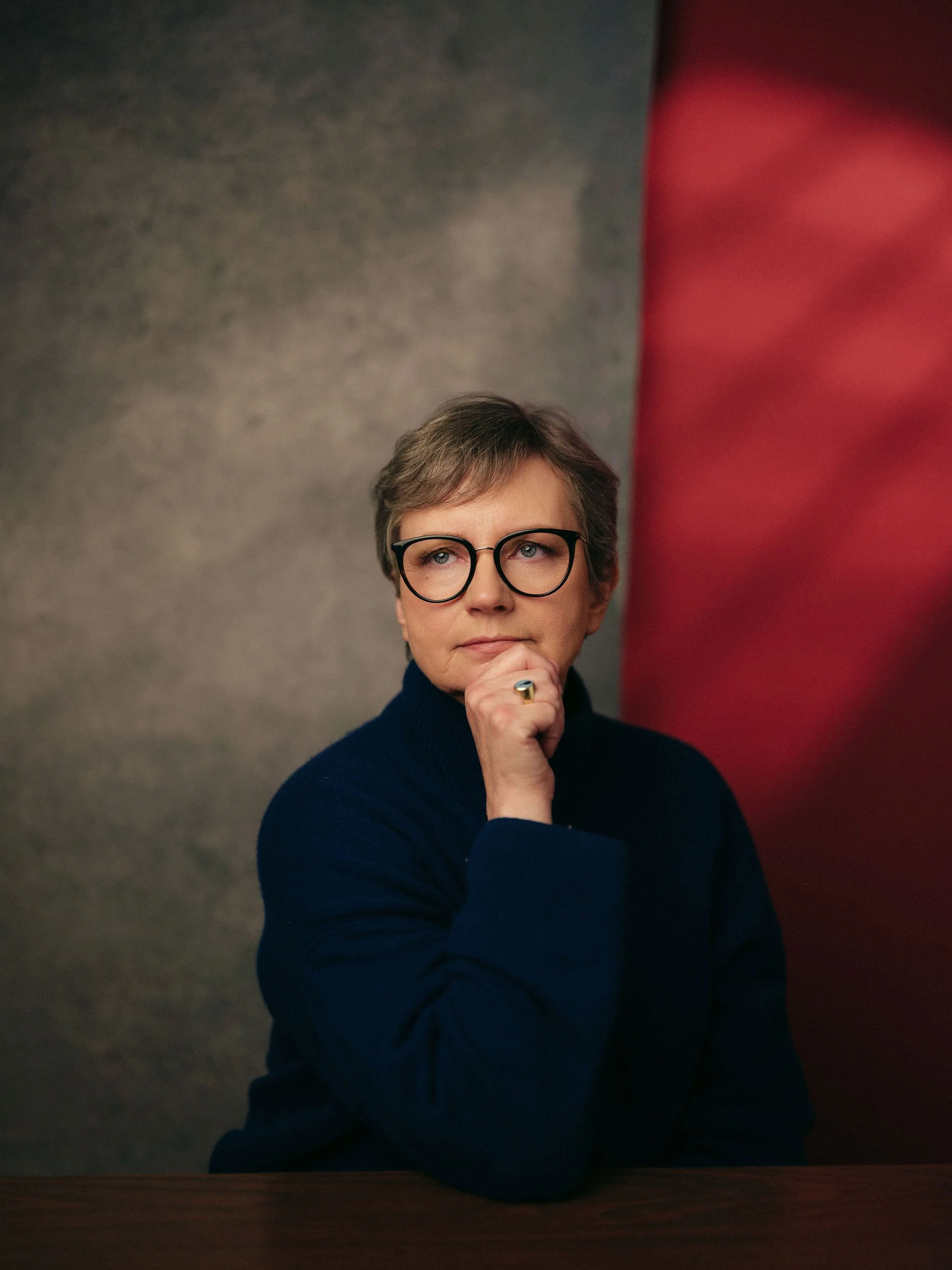 A woman with short gray hair and glasses, wearing a dark sweater, poses with her hand on her chin in front of a textured gray wall and a red wall.