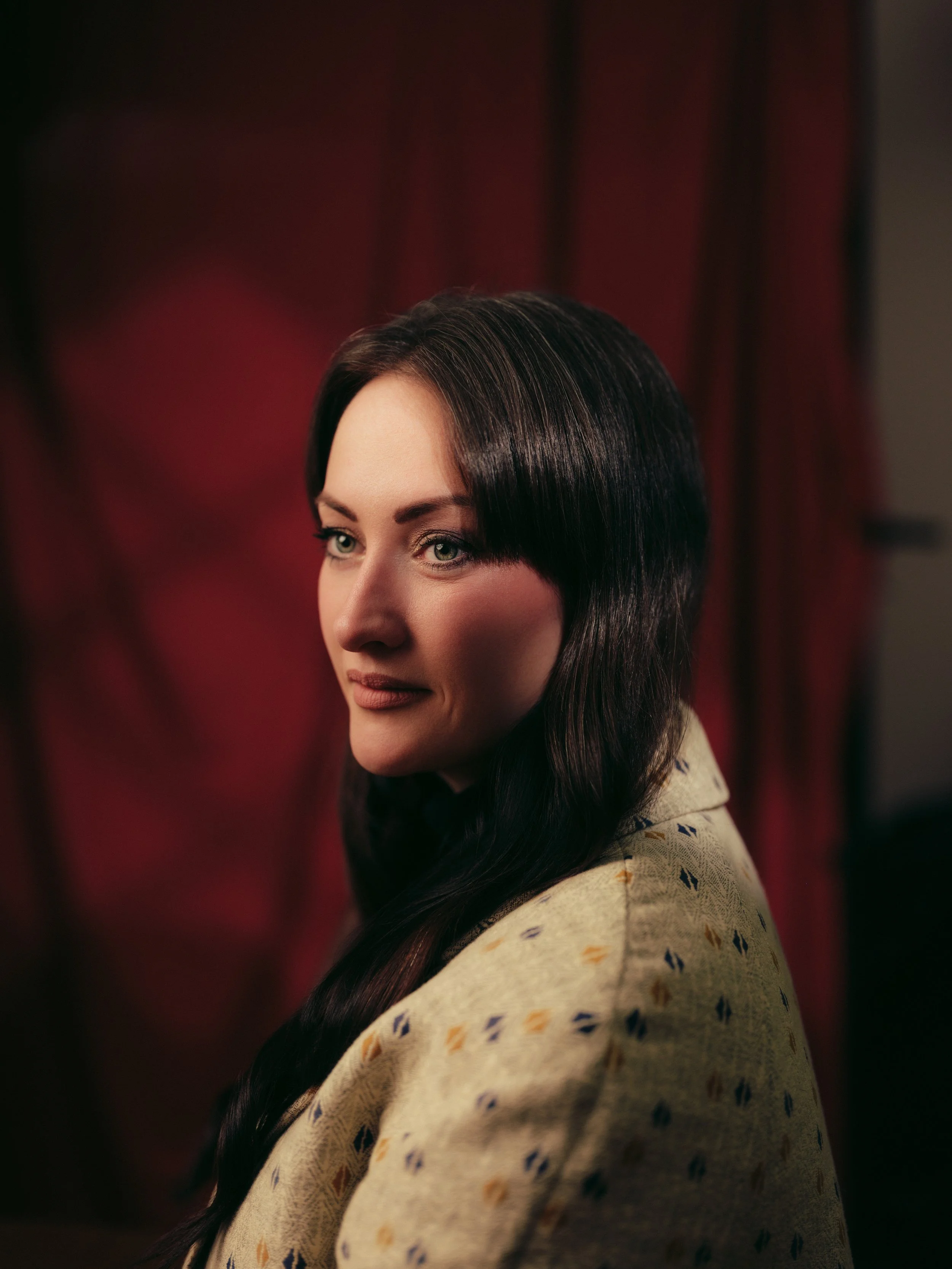 A woman with long dark hair and fair skin looking to her left, wearing a beige sweater with colorful patterns, standing in front of a red curtain.