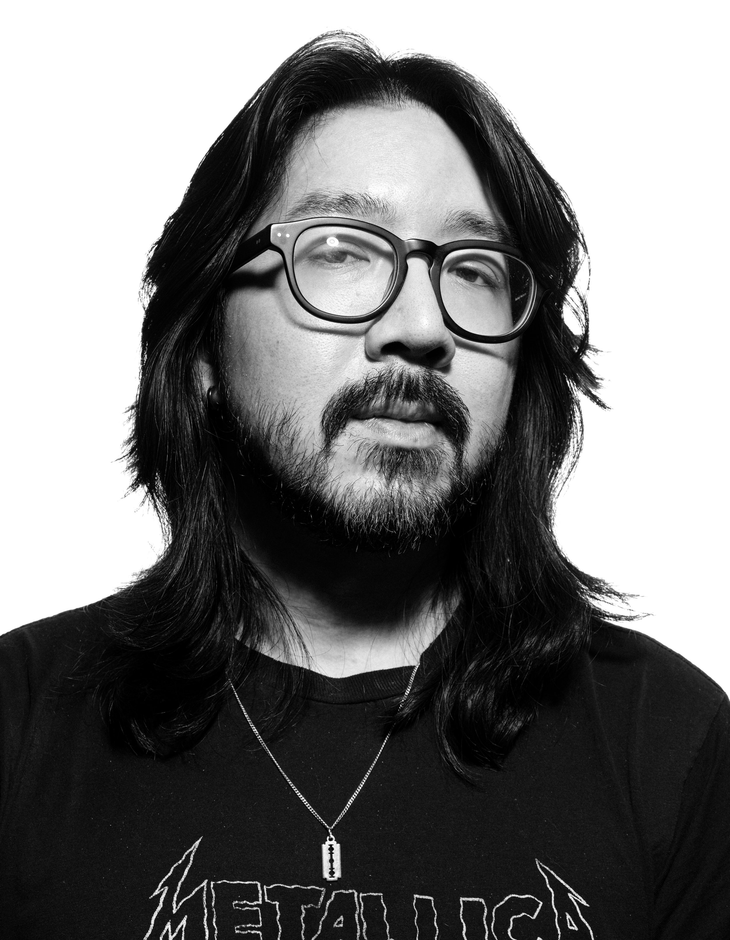 Black and white photo of a man with long dark hair, glasses, and a beard, wearing a black t-shirt with graphic text and a zipper pendant necklace.