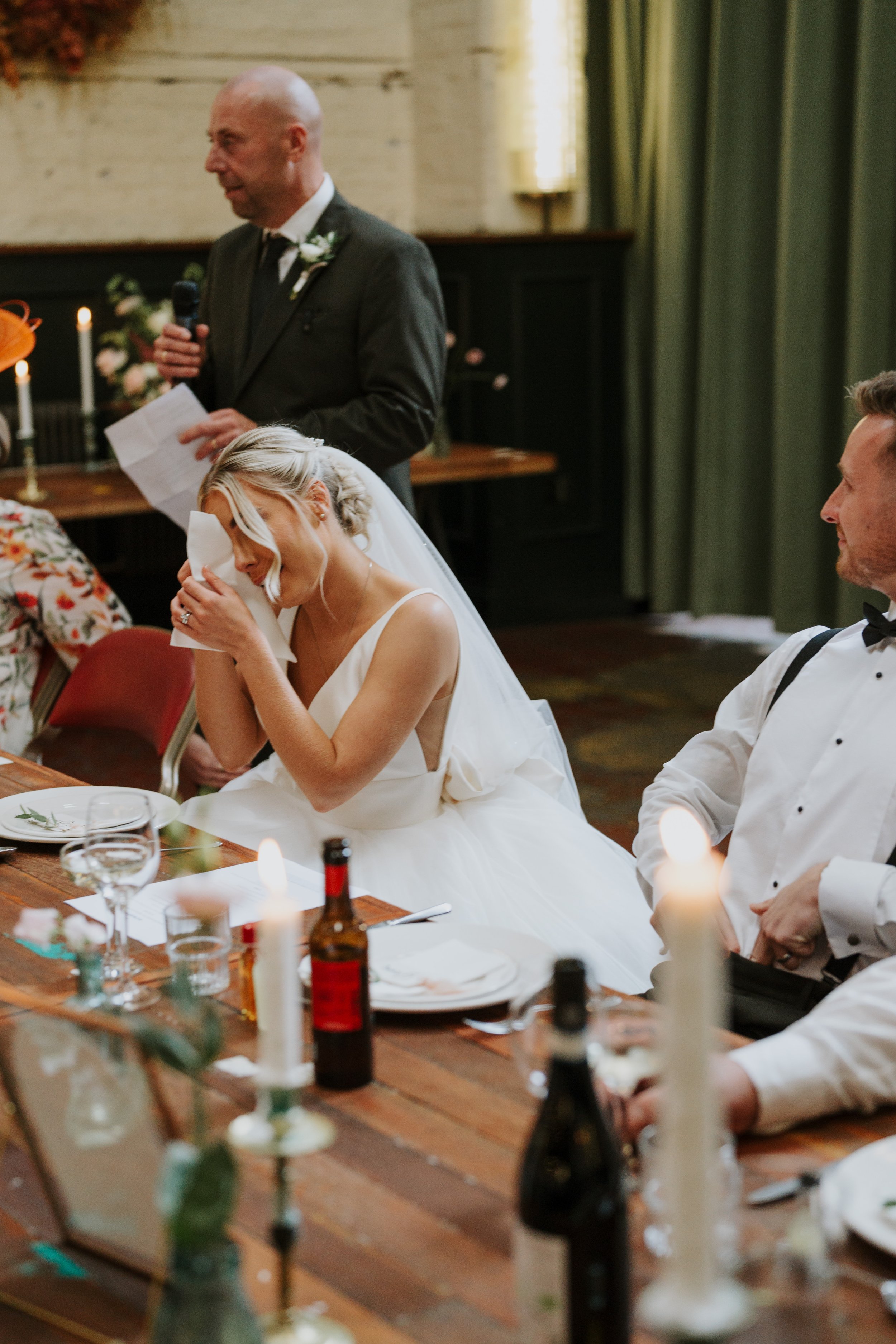 Bride wiping tears, emotional groom and speaker at wedding reception.