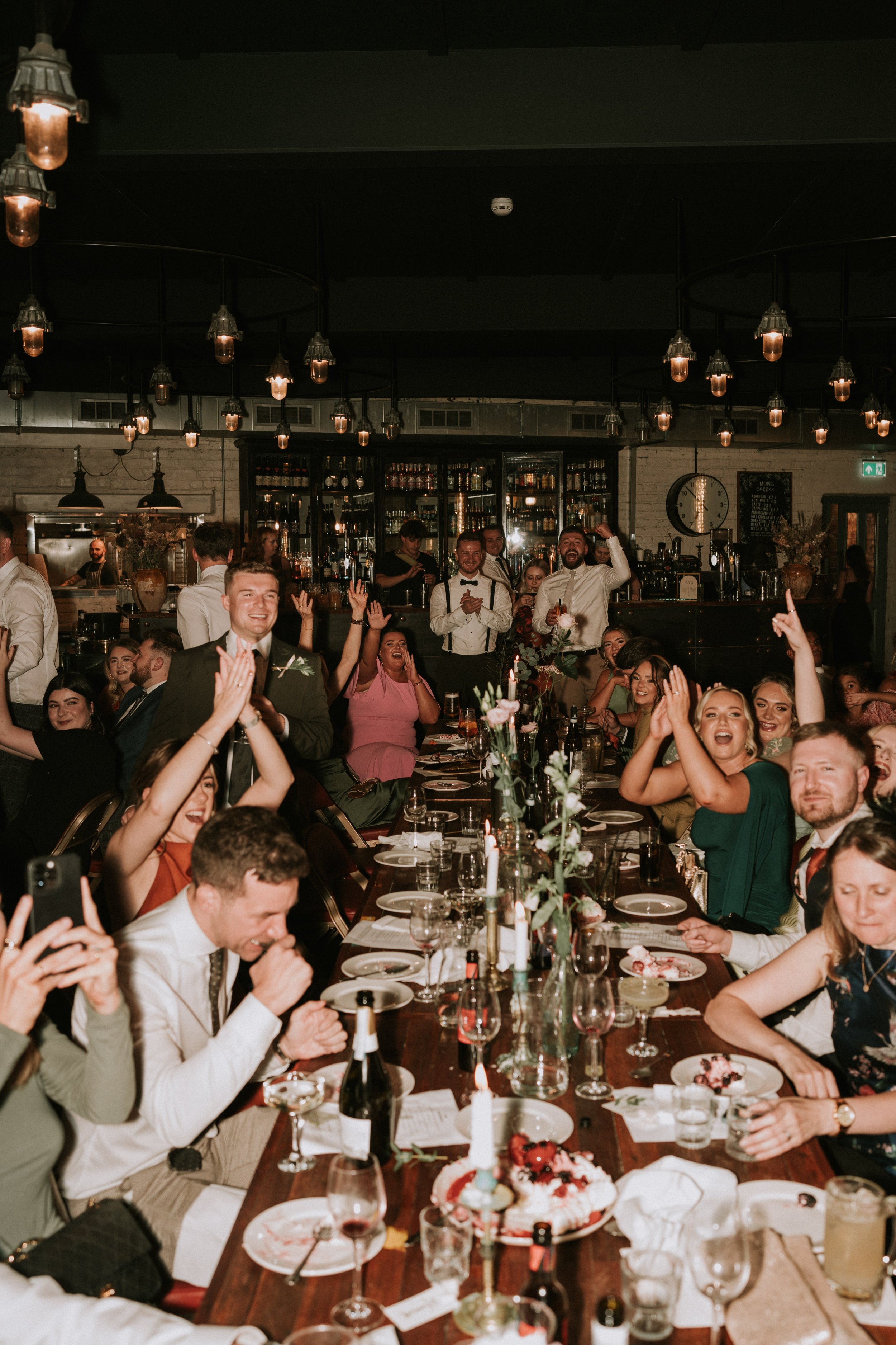 People celebrating at a restaurant or event venue, seated around a long table with food and drinks, under warm lighting.