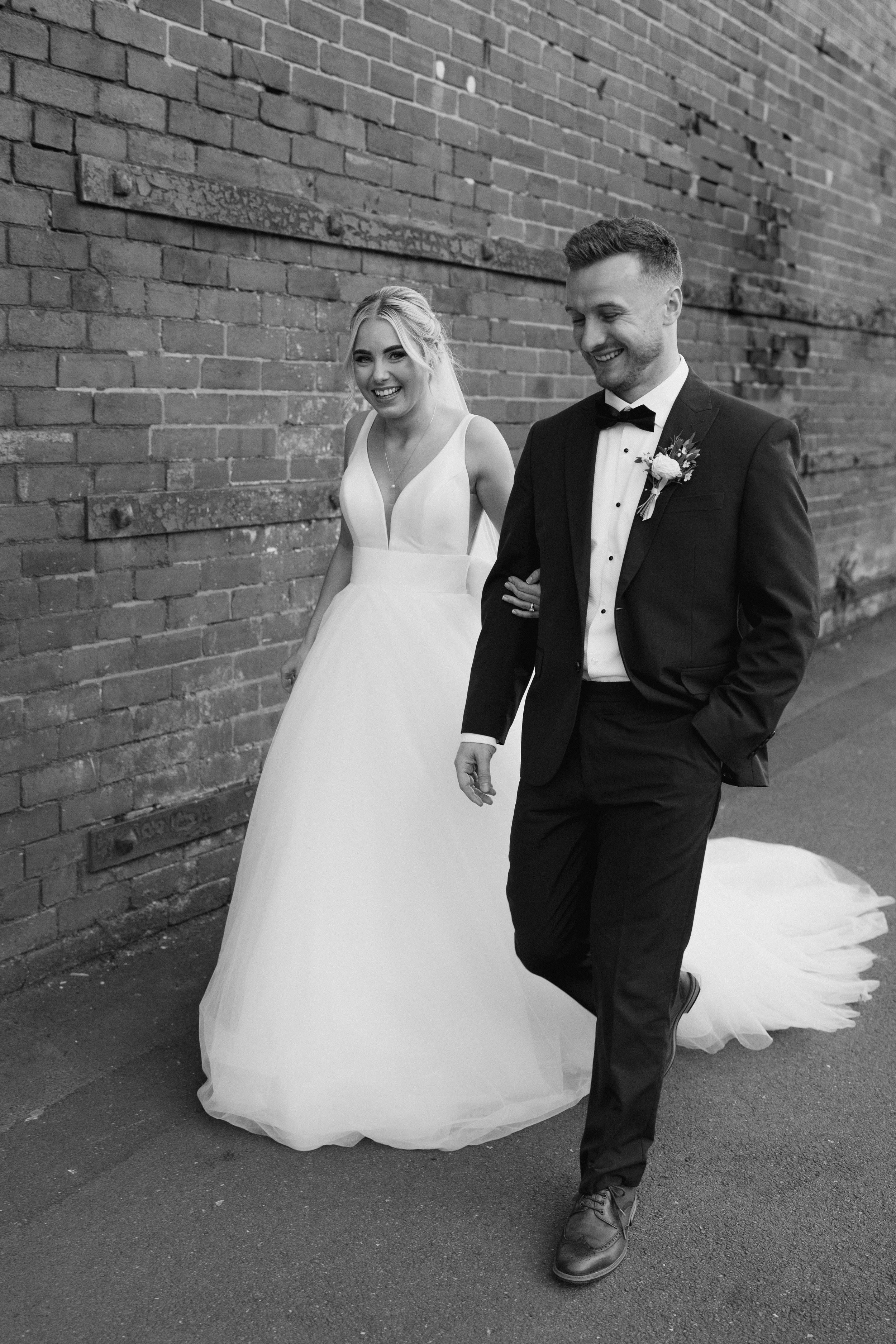 Bride and groom walking beside a brick wall, bride in white gown, groom in black tuxedo.