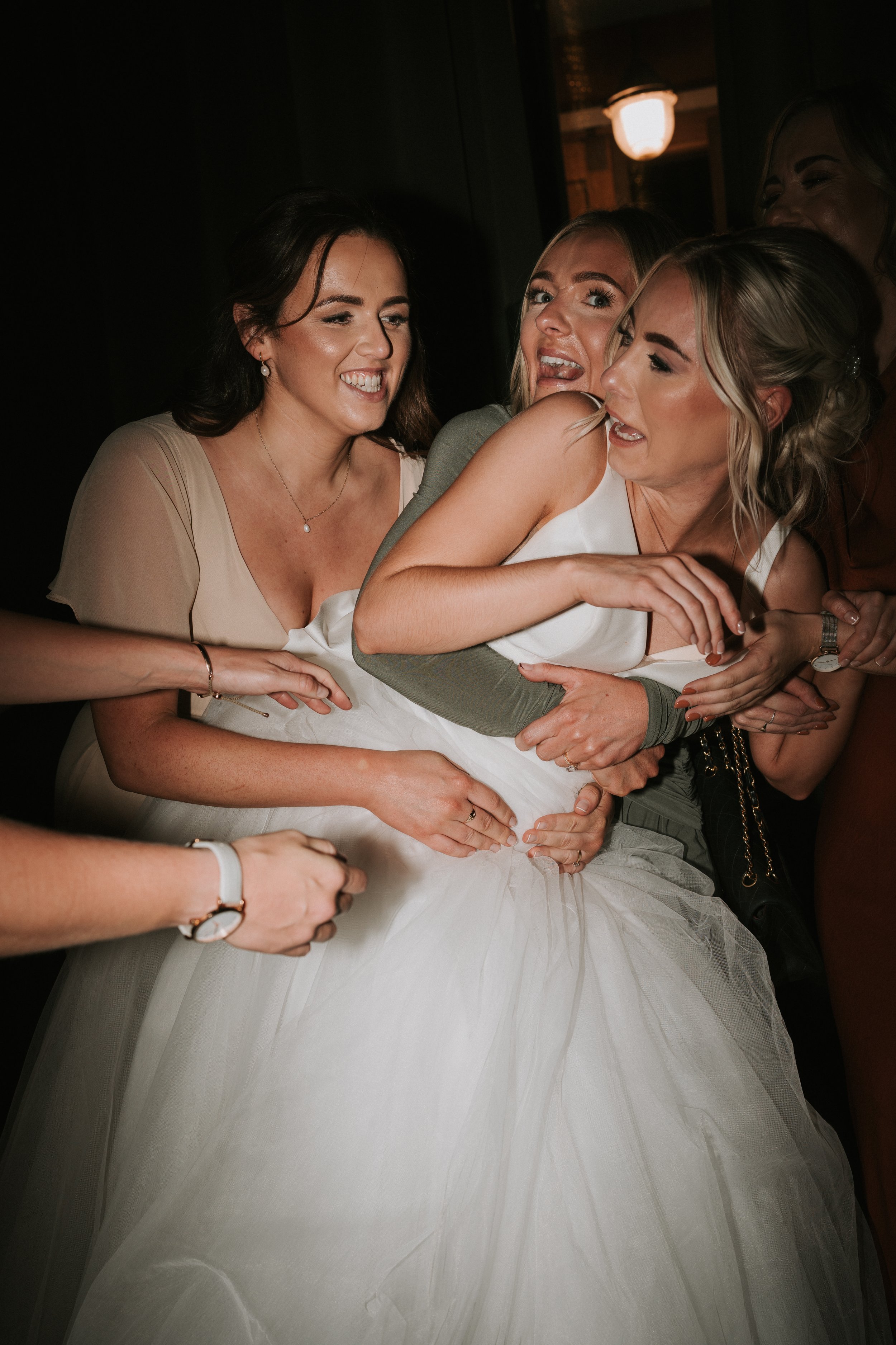 Group of women embracing and laughing, one in a wedding dress, in a lively, candid moment.