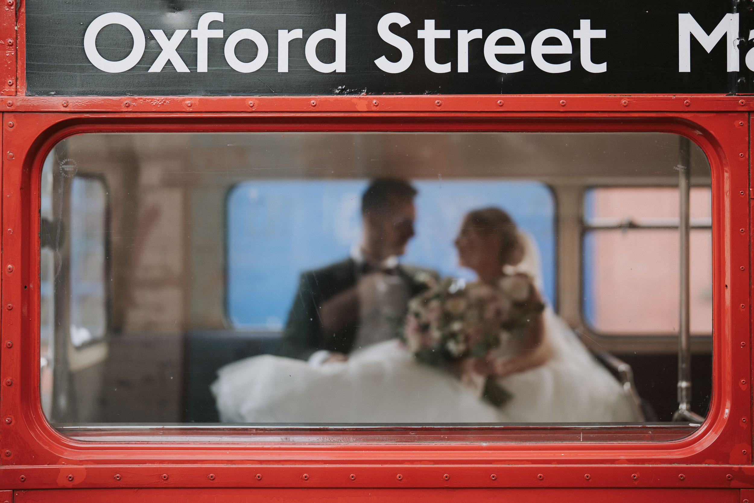 Couple inside a red double-decker bus, partially obscured lettering 'Oxford Street', bride holding a bouquet.