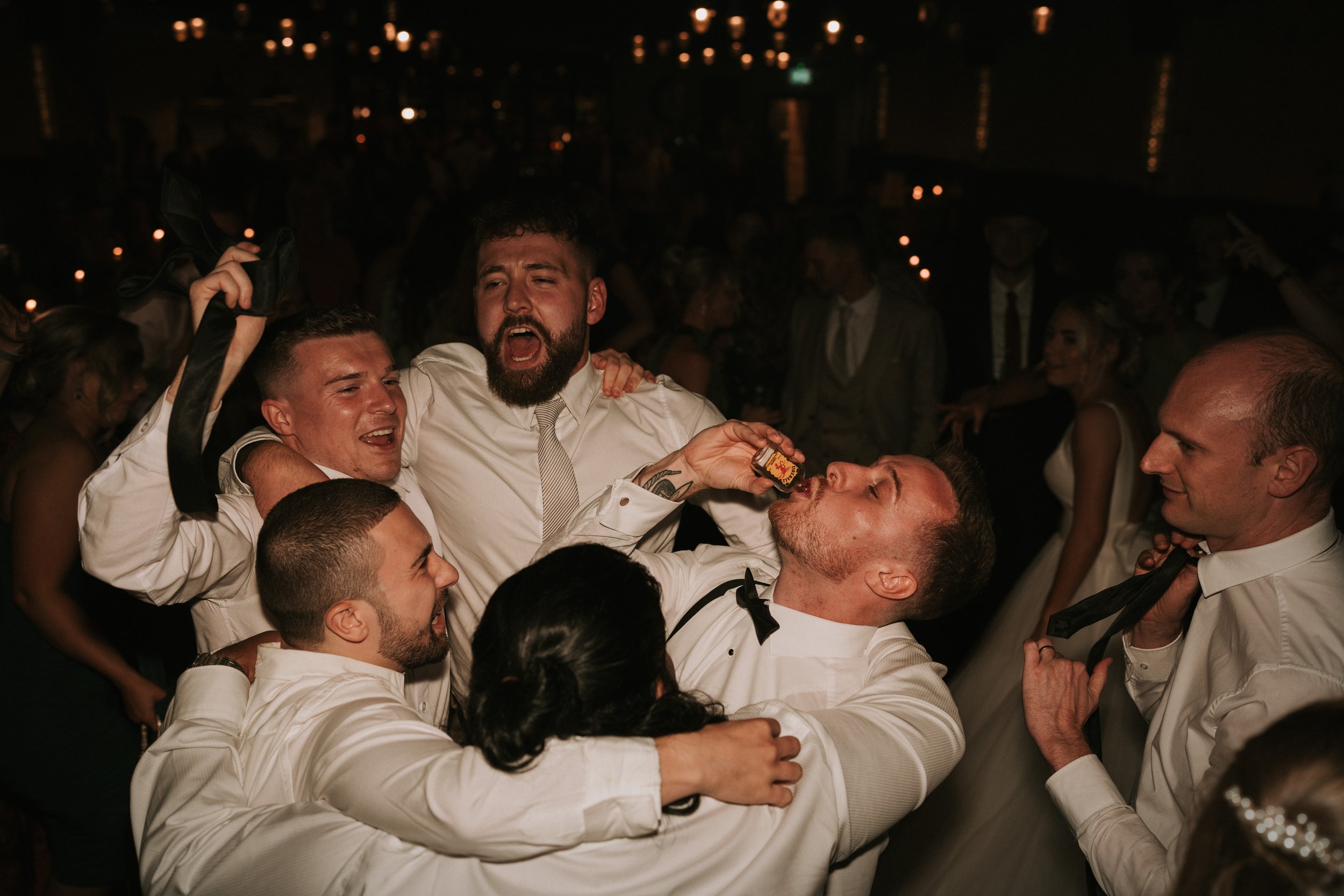 Group of people celebrating at a party, one person drinking a shot, wearing formal attire, dark ambiance with blurred background.