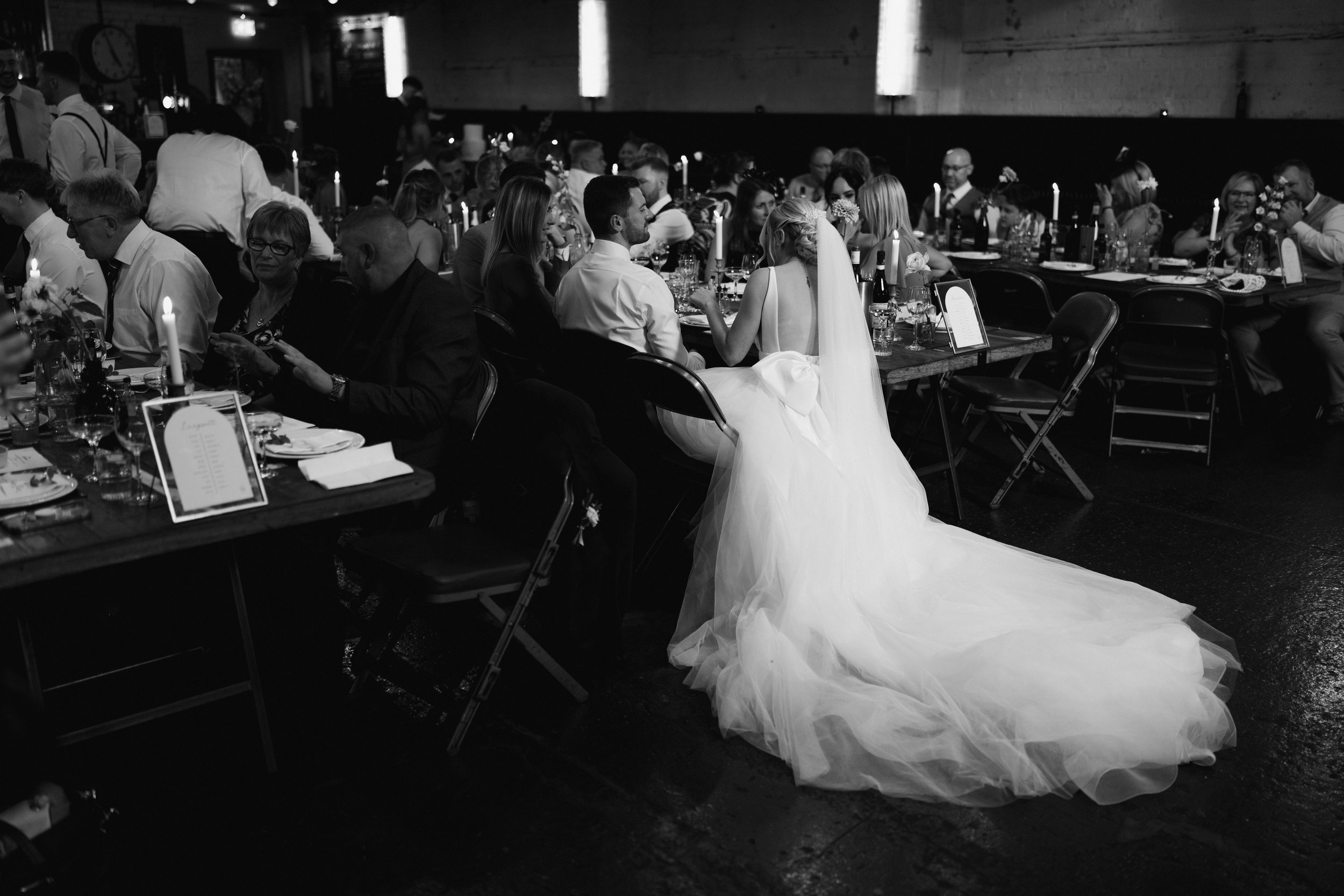 Black and white wedding reception scene with bride in a long train dress seated at a banquet table among guests.