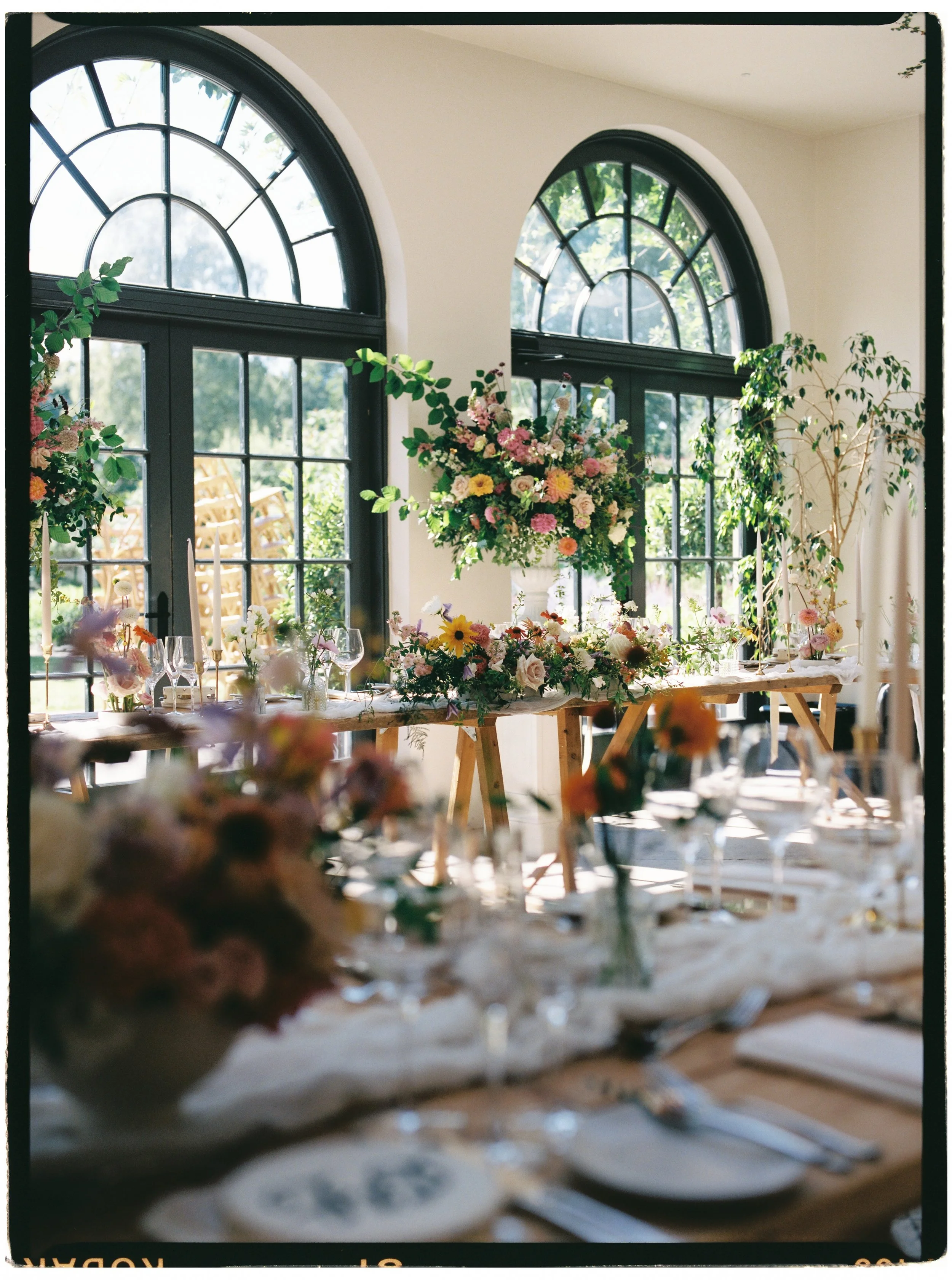 An indoor event space decorated with floral arrangements, large arched windows, and a table set for a formal gathering.