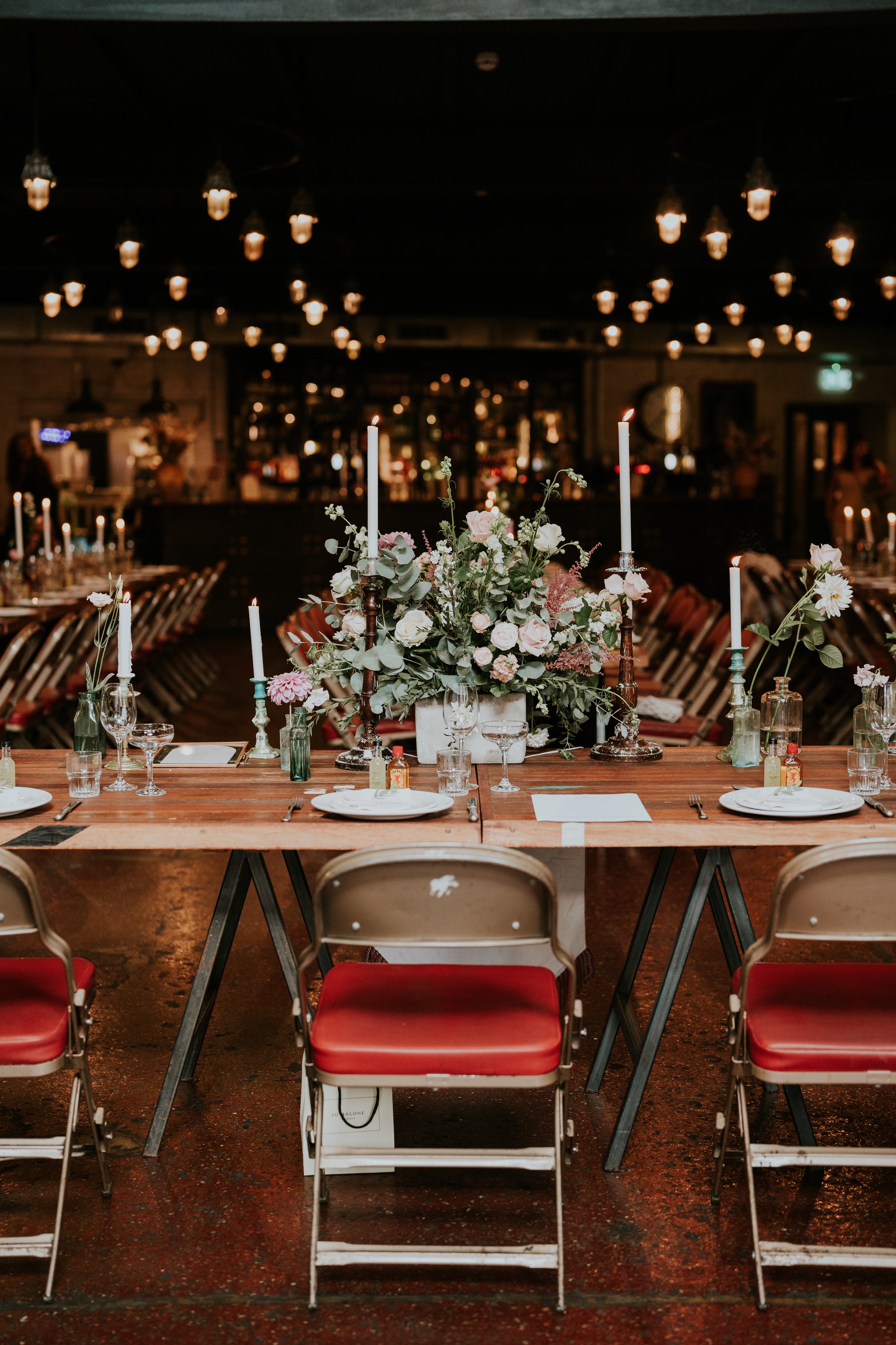 Elegant dining table setup with flowers and candles in a dimly lit venue, featuring vintage decor and overhead lights.