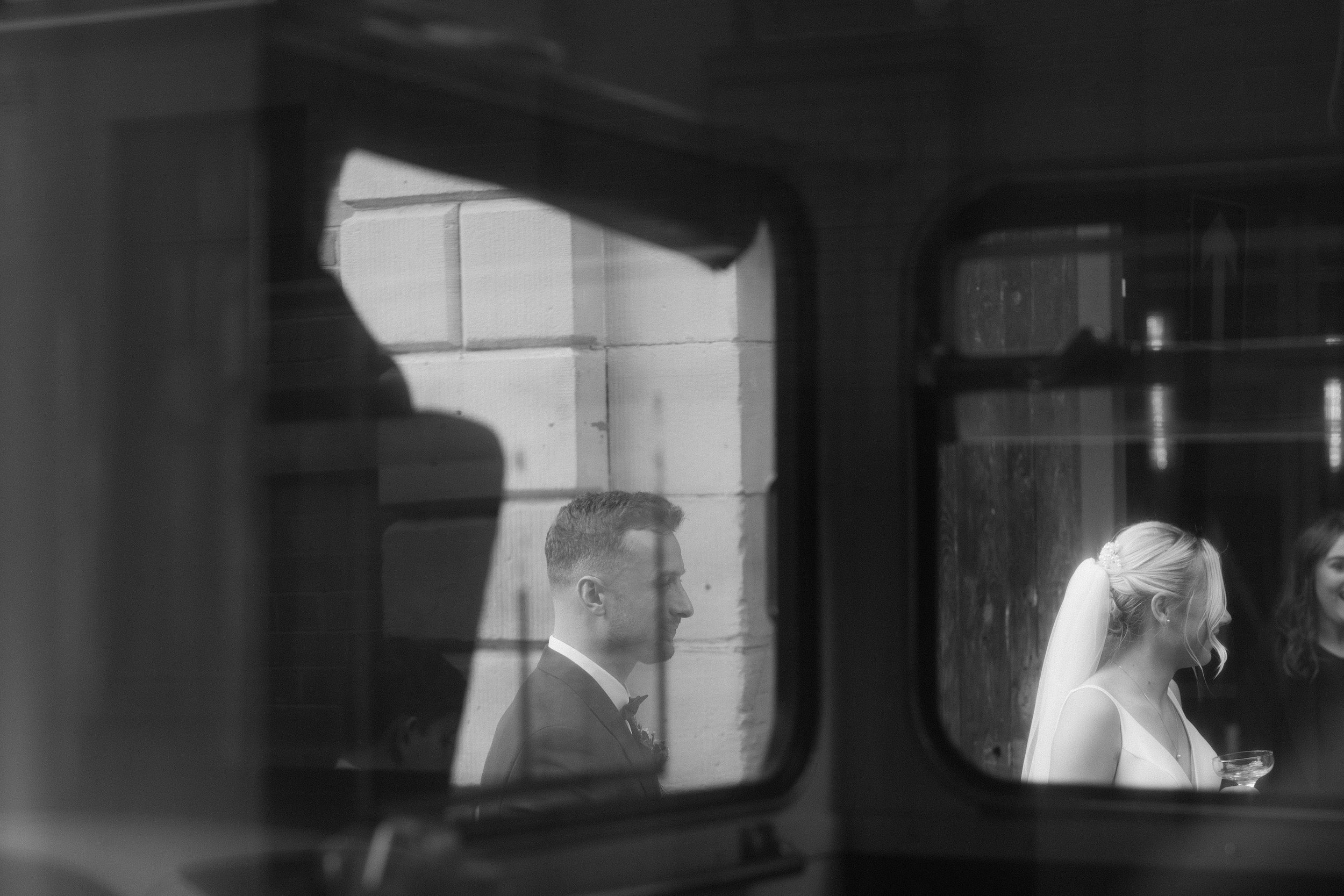 Black and white image of a bride and groom seen through a window reflection. The bride is holding a glass, wearing a veil.