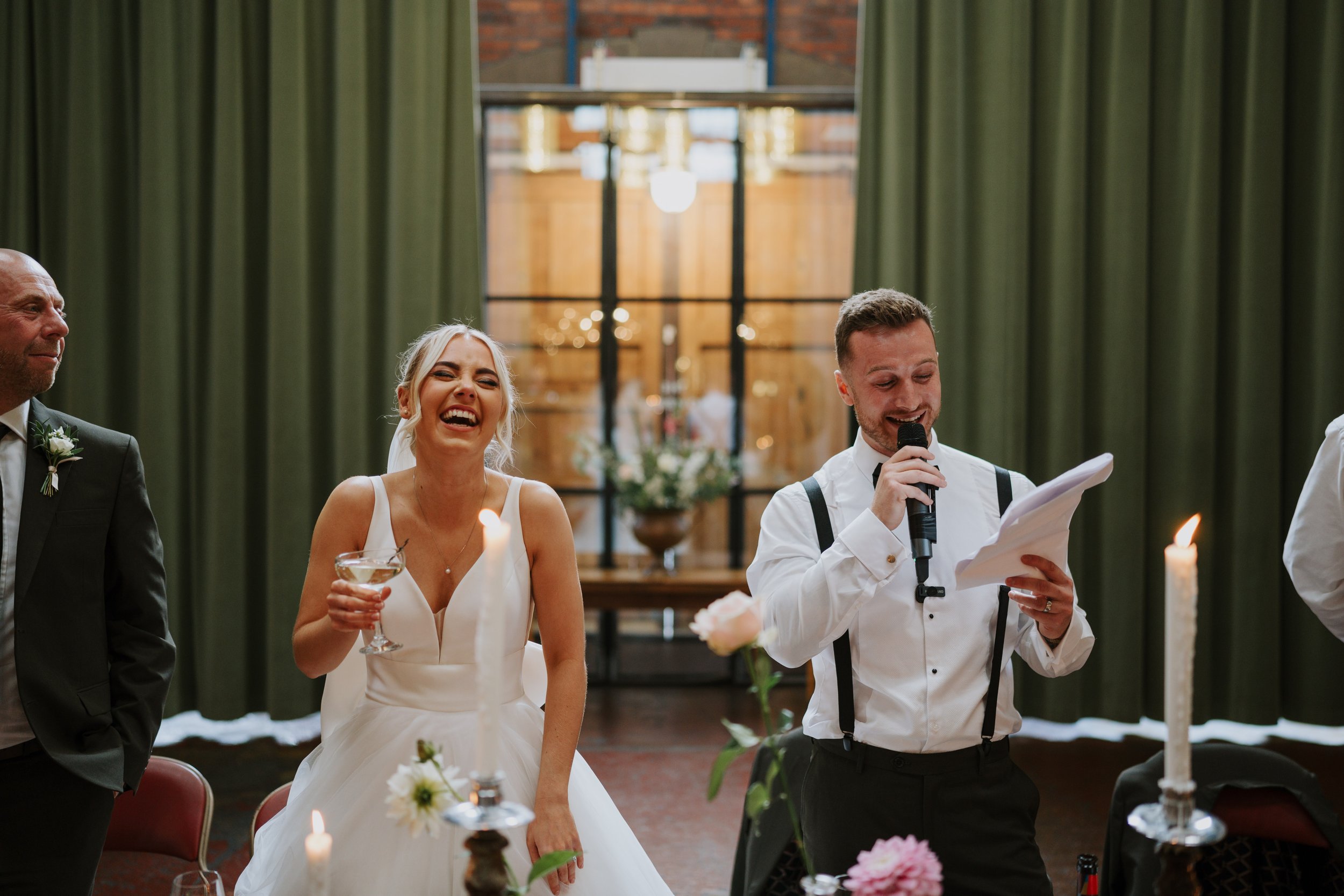 Bride laughing with guests during wedding speech, indoor setting, candles and flowers on table.