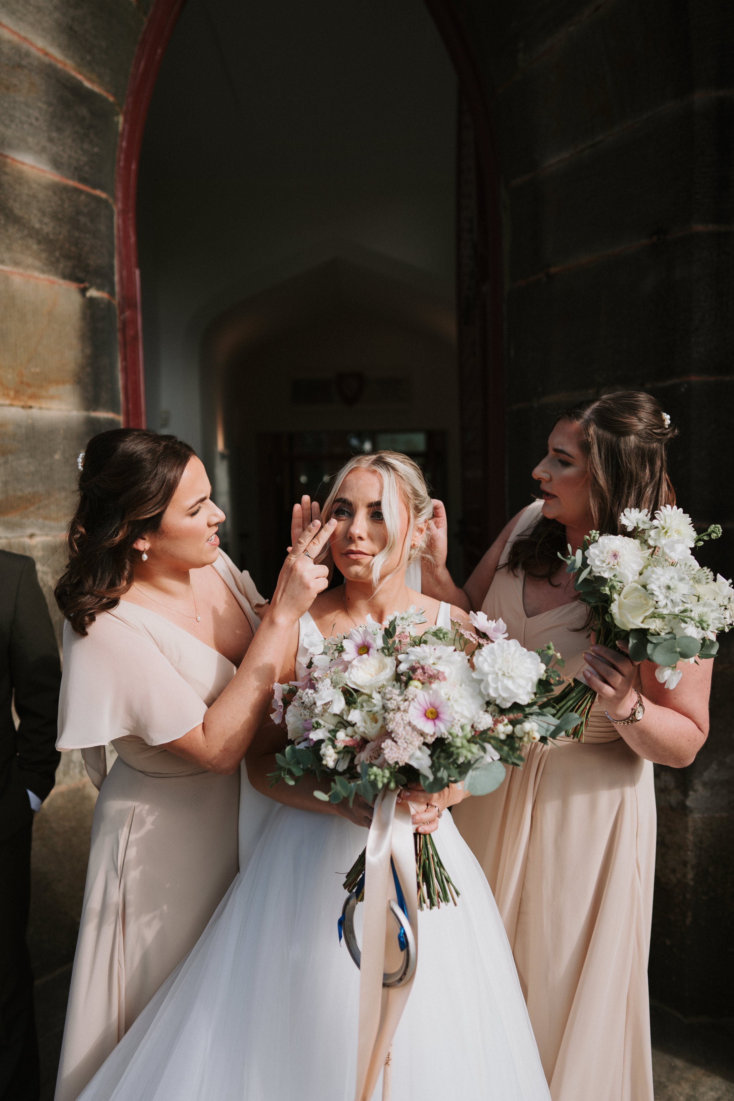 Bride holding a bouquet with two bridesmaids adjusting her hair outside a building.