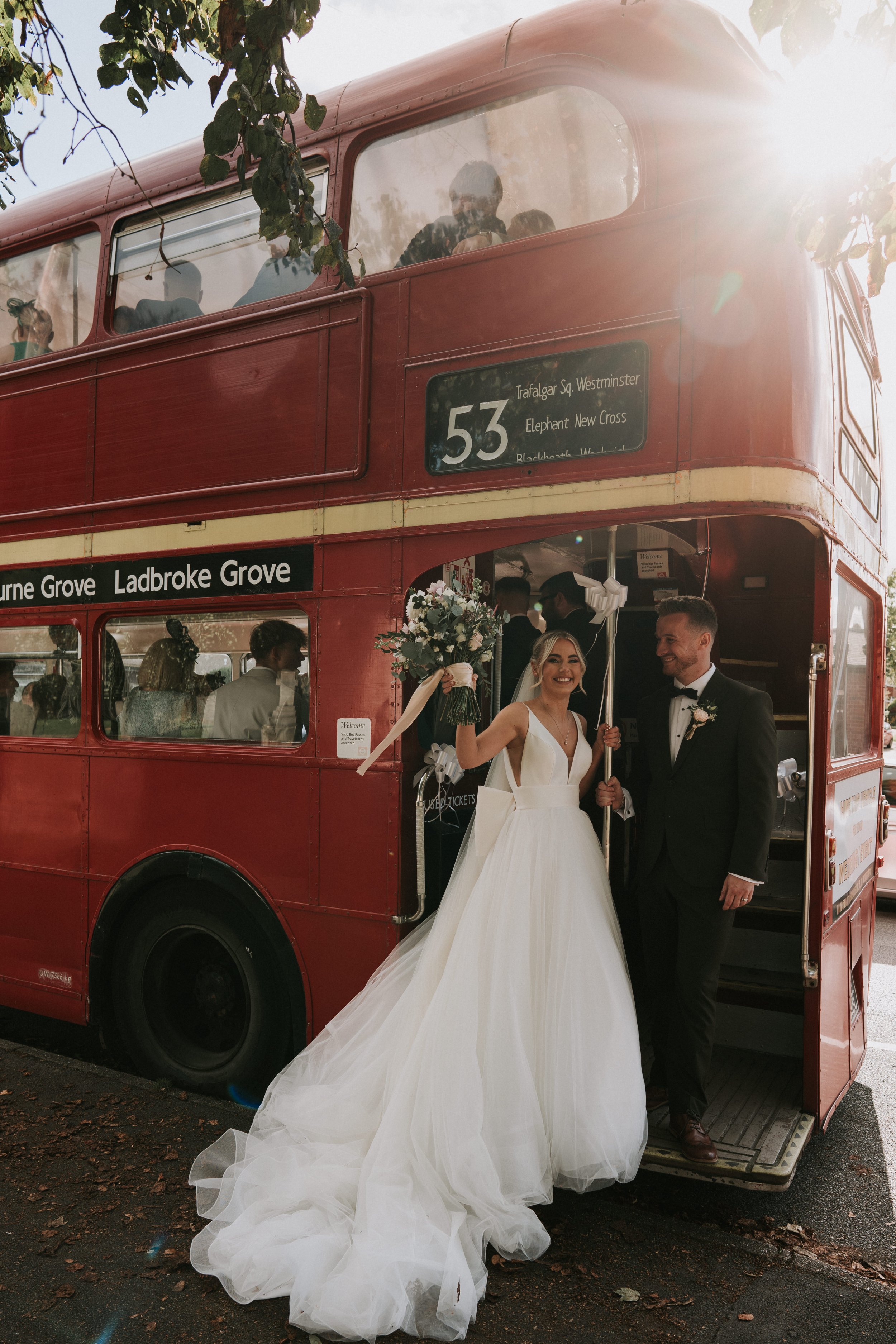 Bride and groom standing by a vintage red double-decker bus, with the bride holding a bouquet and wearing a white wedding dress.