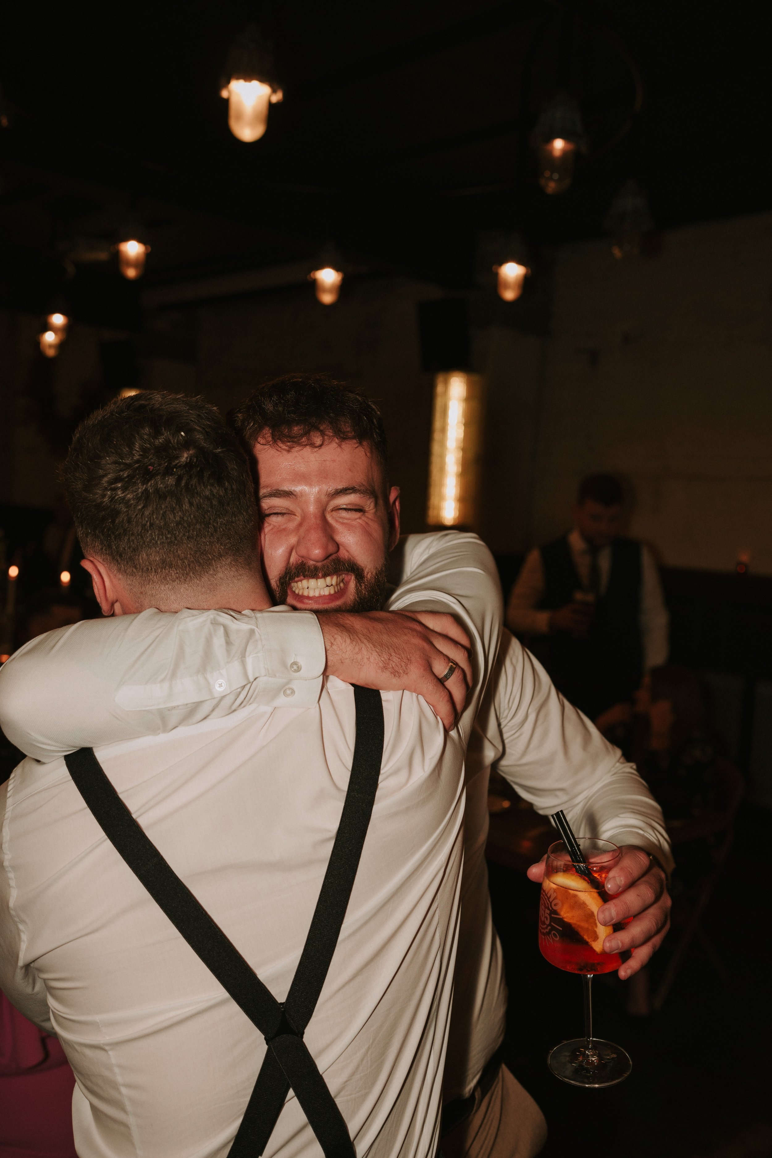 Two men in white shirts hugging at a party, one holding a cocktail with orange slice and straw, warmly lit atmosphere with vintage lights.