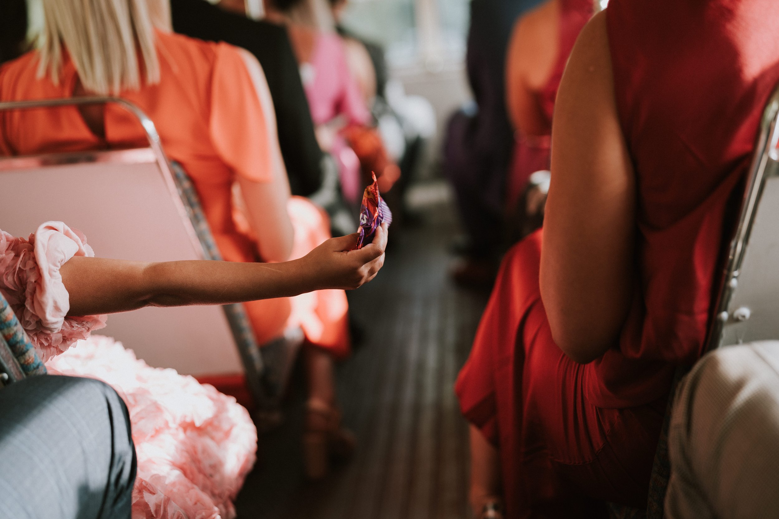 People sitting on a bus in colorful clothing, with a focus on a hand holding a packaged snack.
