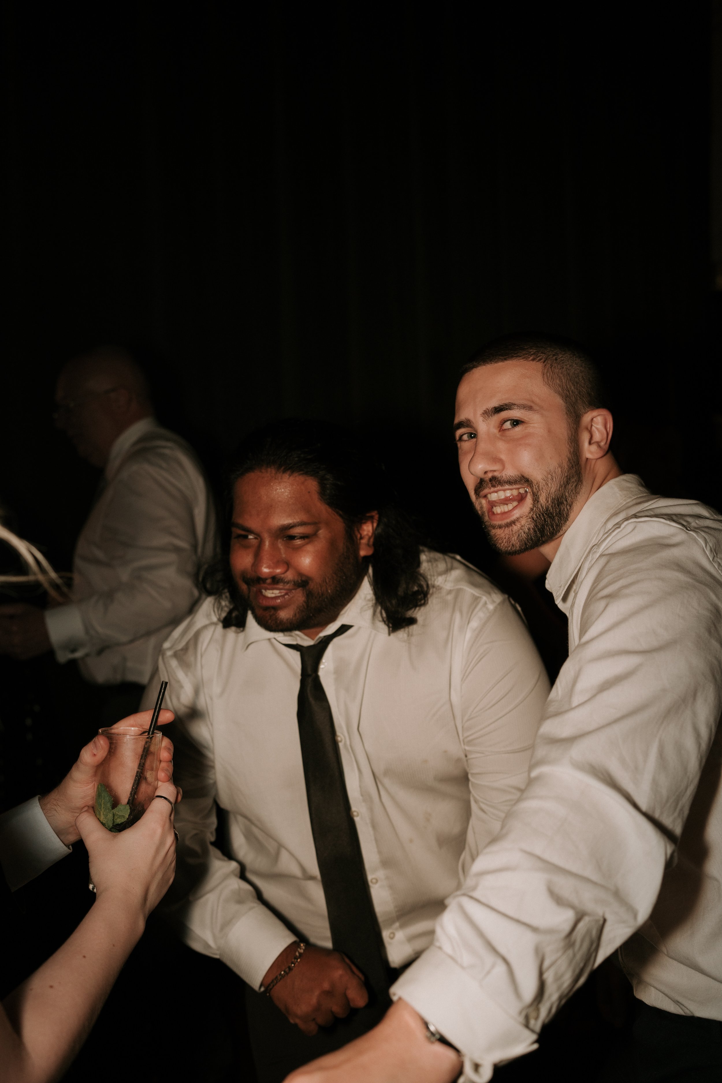 Two men in formal attire smiling at a party or event, with a third person holding a drink in the foreground.