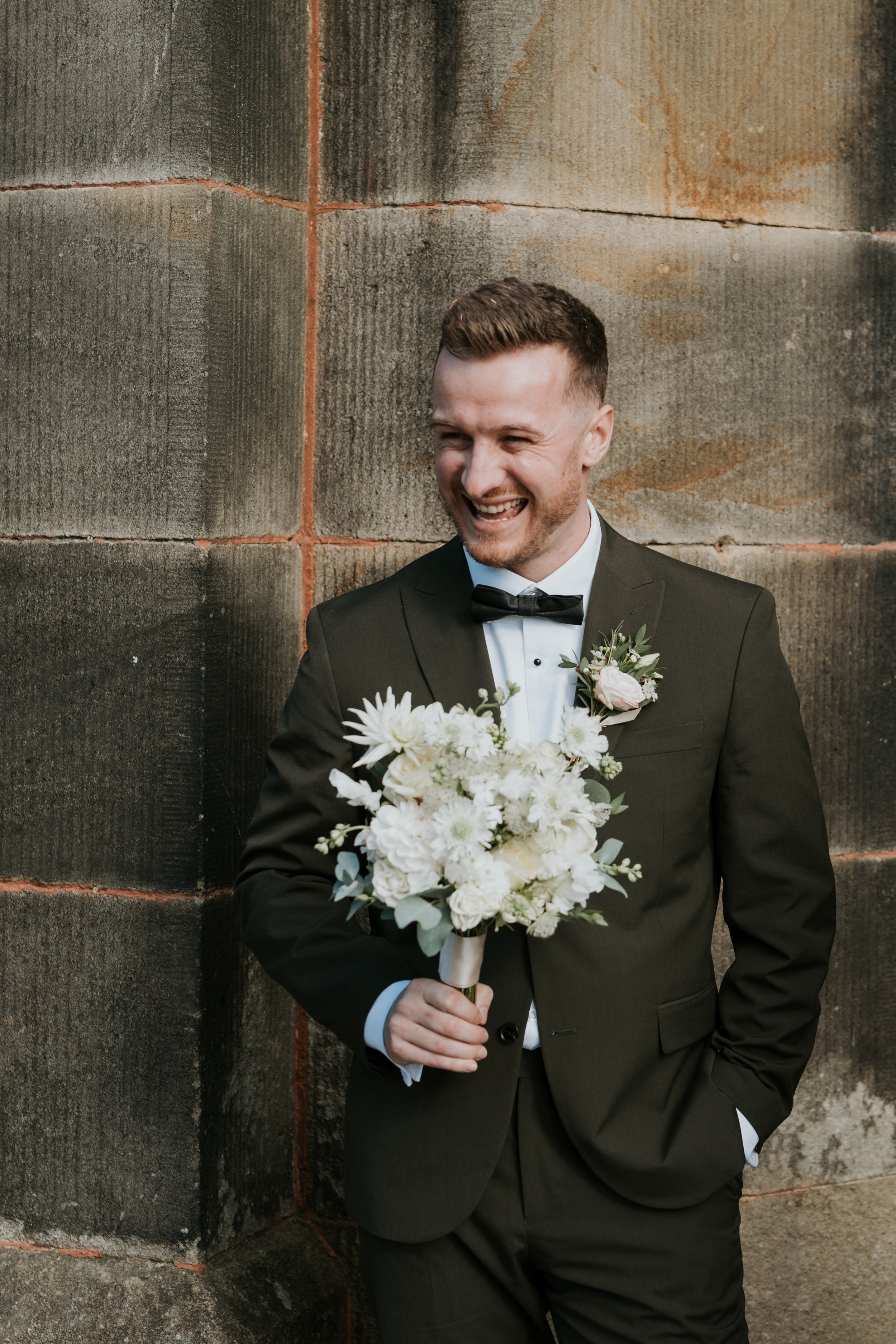 Man in suit holding bouquet, smiling, standing against stone wall