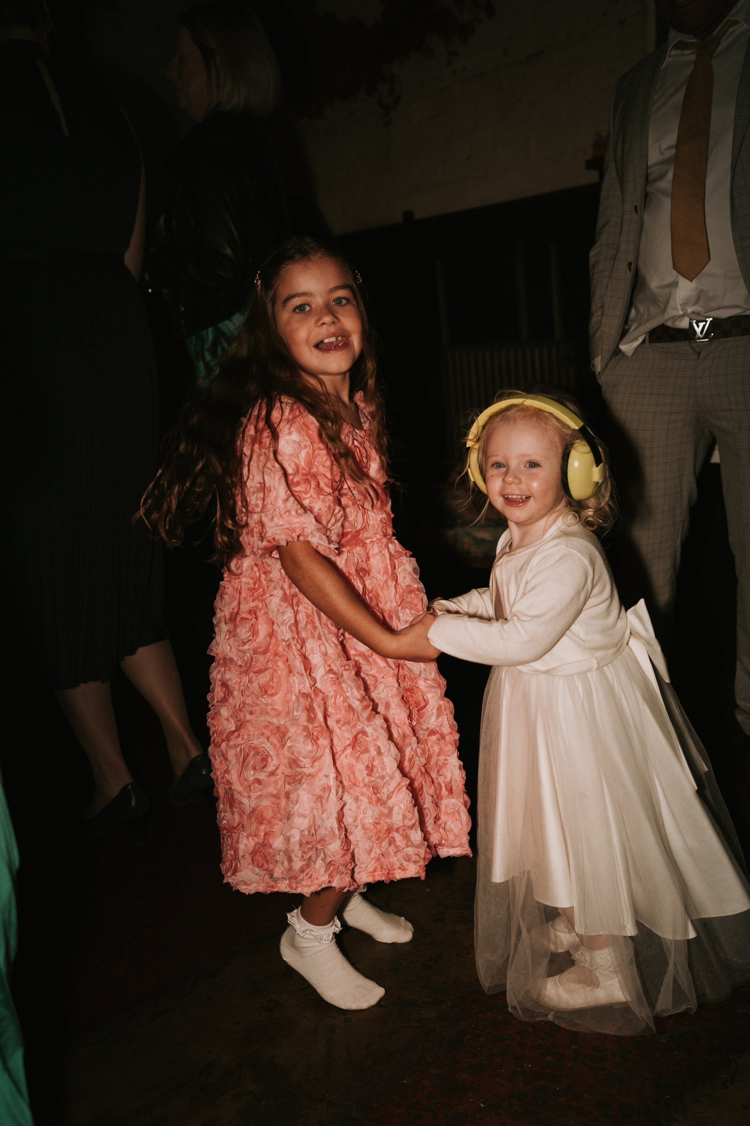 Two young girls smiling and holding hands, one in a pink floral dress and the other in a white dress with headphones, likely at a formal event.