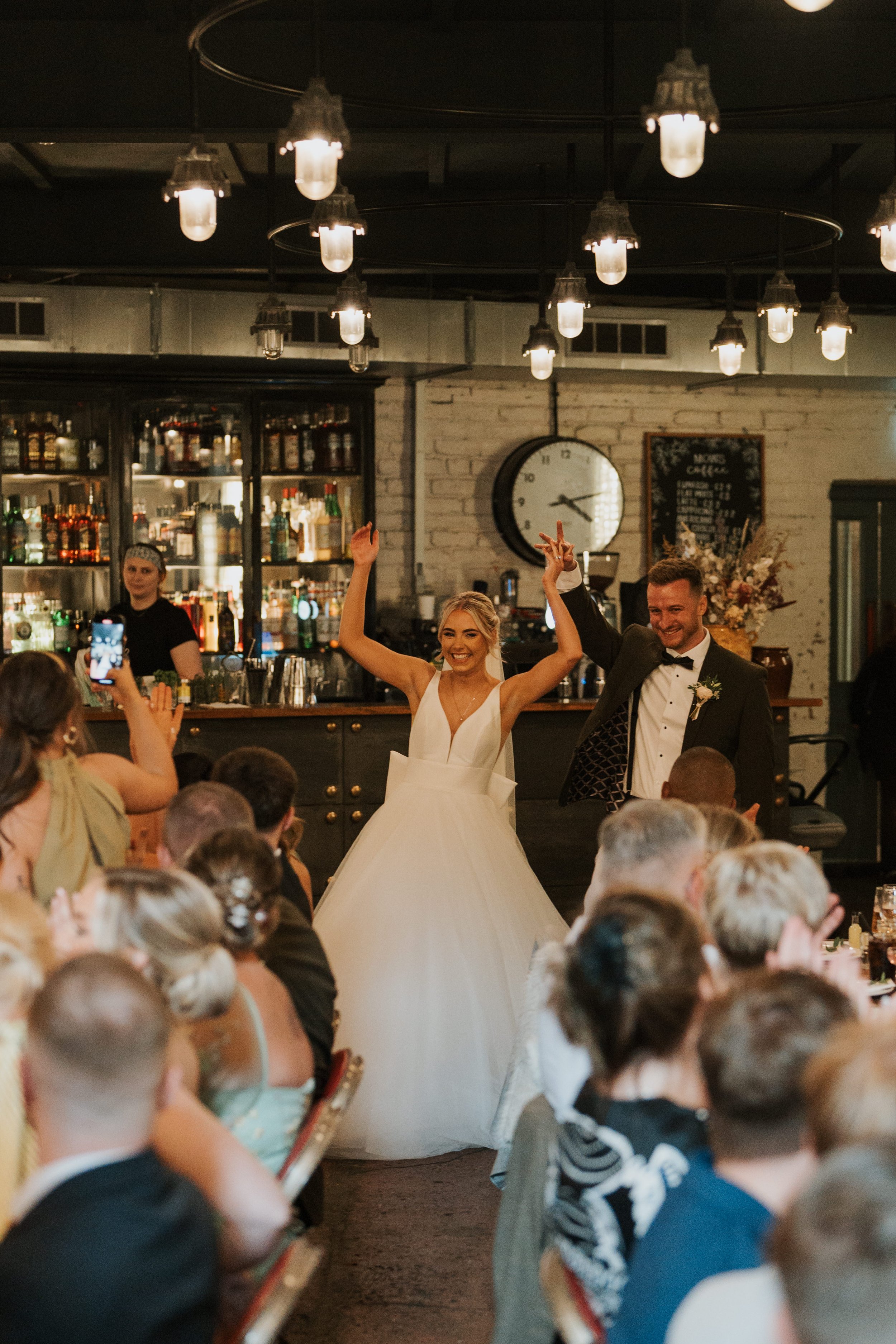 Bride and groom entering a wedding reception, smiling with hands raised, surrounded by seated guests in a warmly lit venue with a bar in the background.