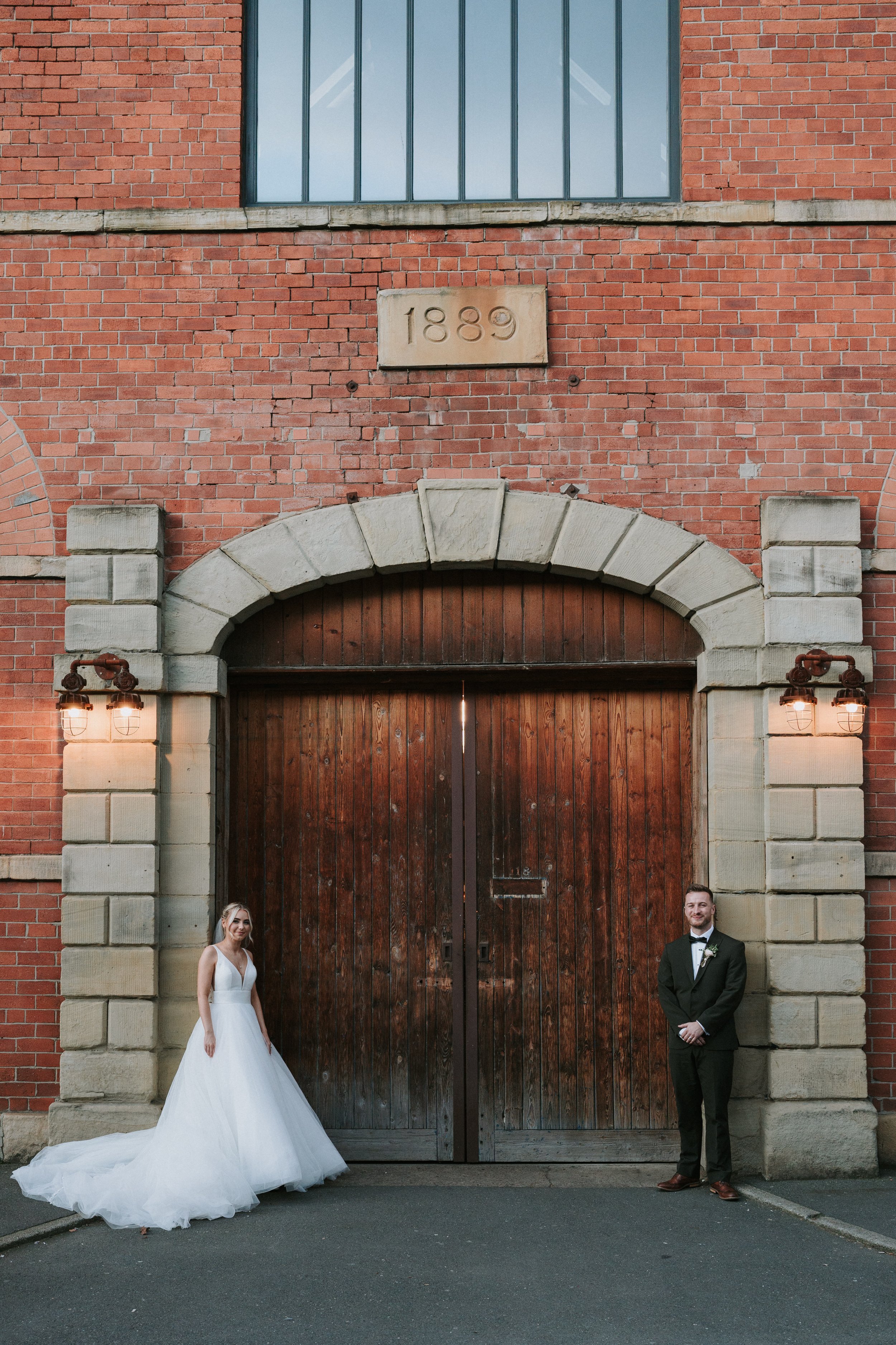 Bride in white gown and groom in suit standing outside brick building with "1889" stone plaque.