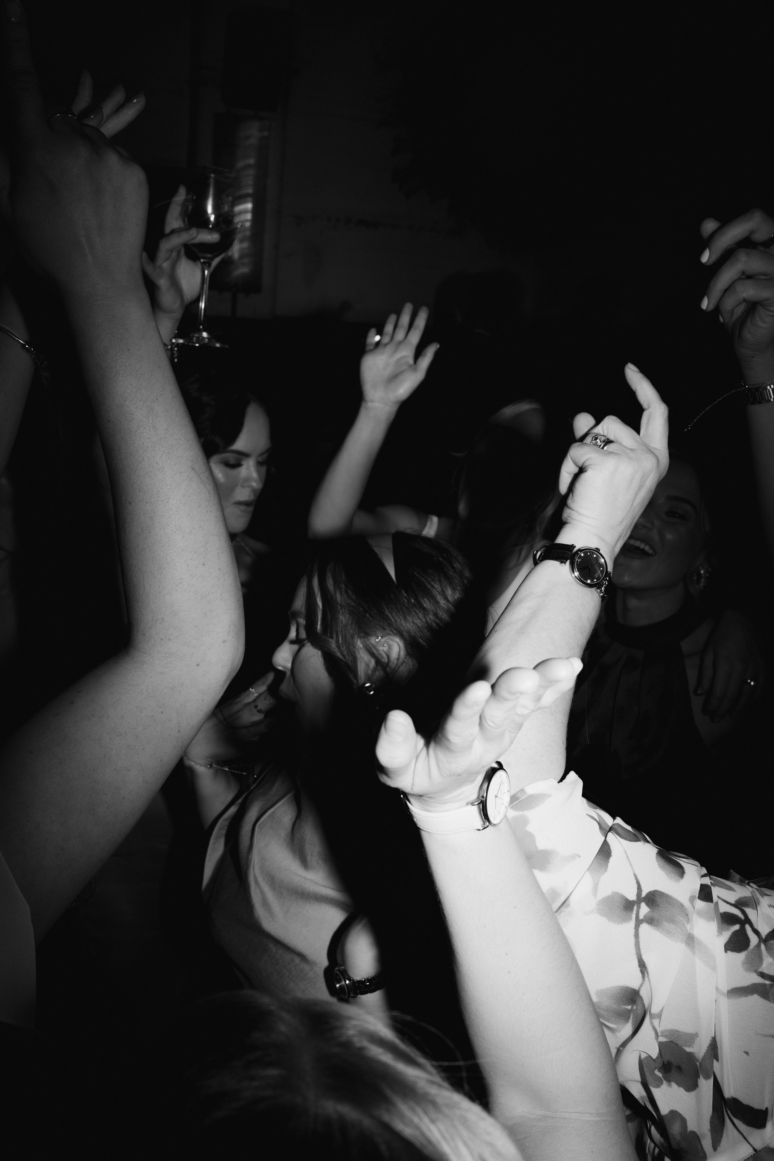 Black and white photo of people dancing at a party, hands raised, holding a wine glass.