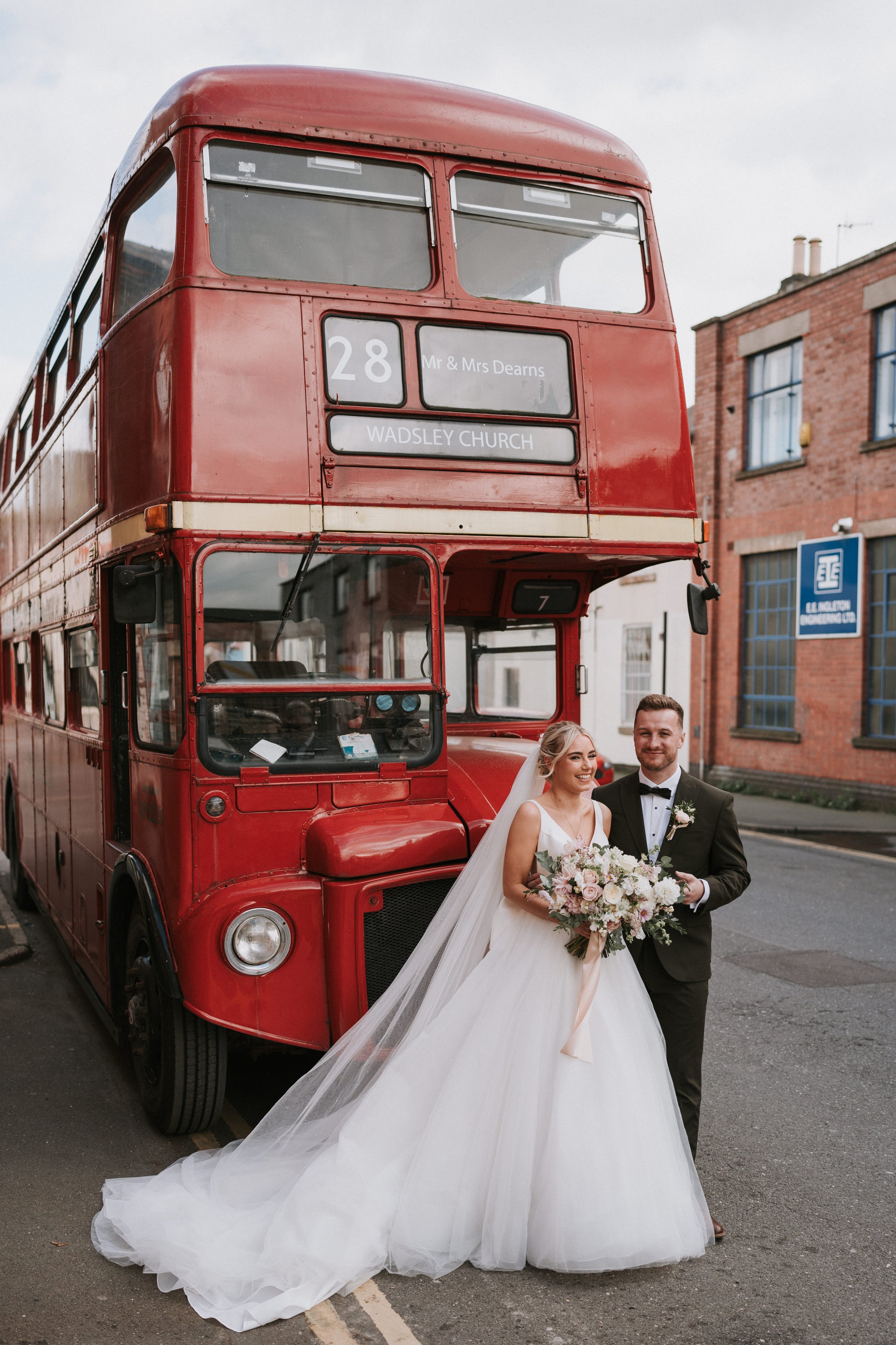 Bride and groom in wedding attire standing in front of a vintage red double-decker bus with "28 Wadsley Church" displayed.